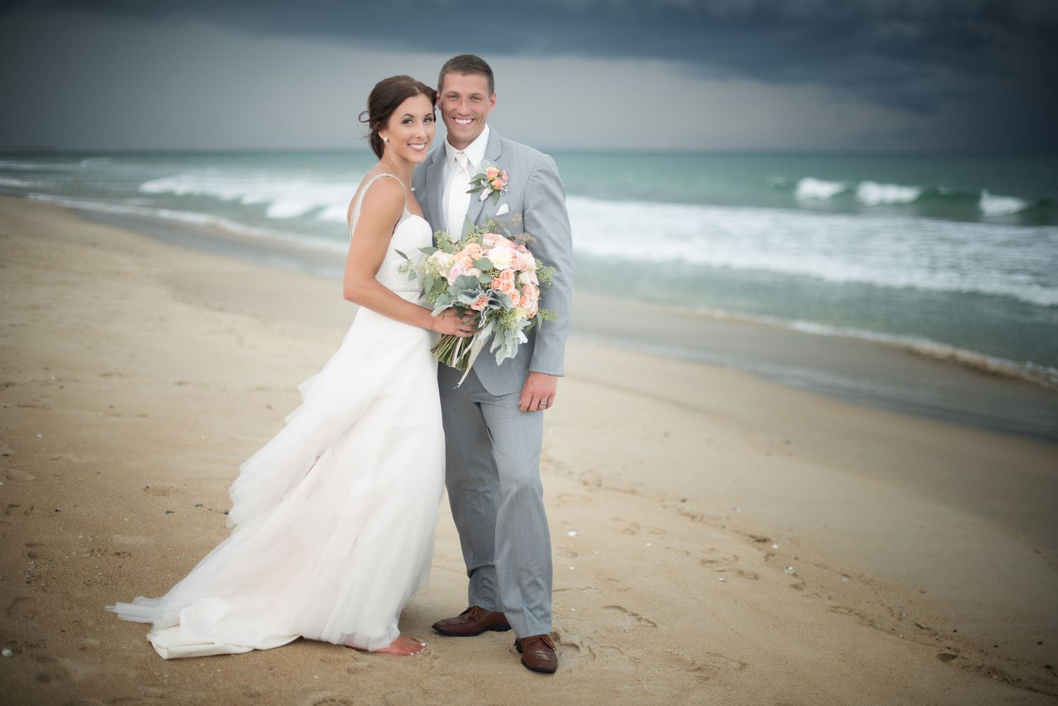 Bride and groom posing together on a sandy beach with stormy ocean waves in the background on their wedding day.