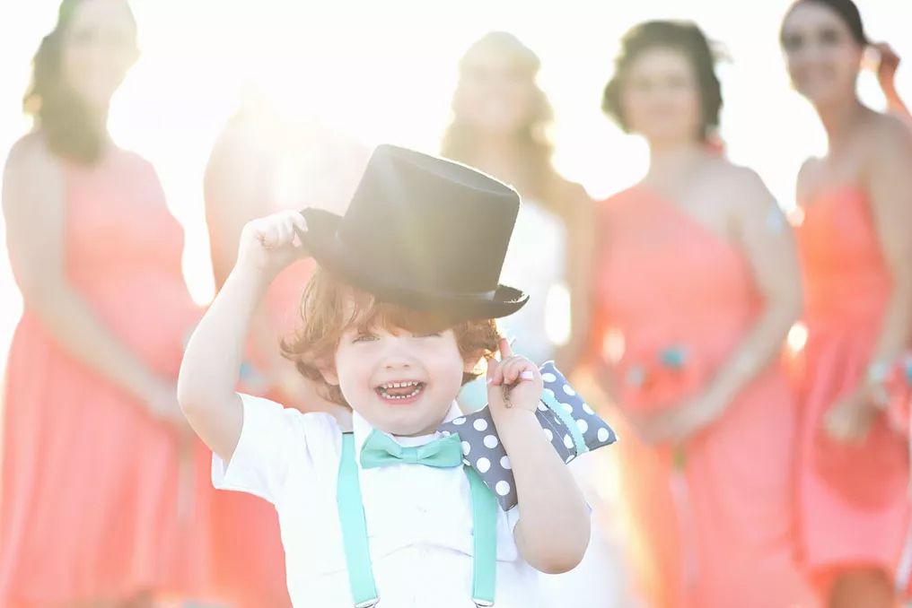 Adorable ring bearer in top hat and bow tie laughs joyfully with coral-dressed bridesmaids in sunny background.
