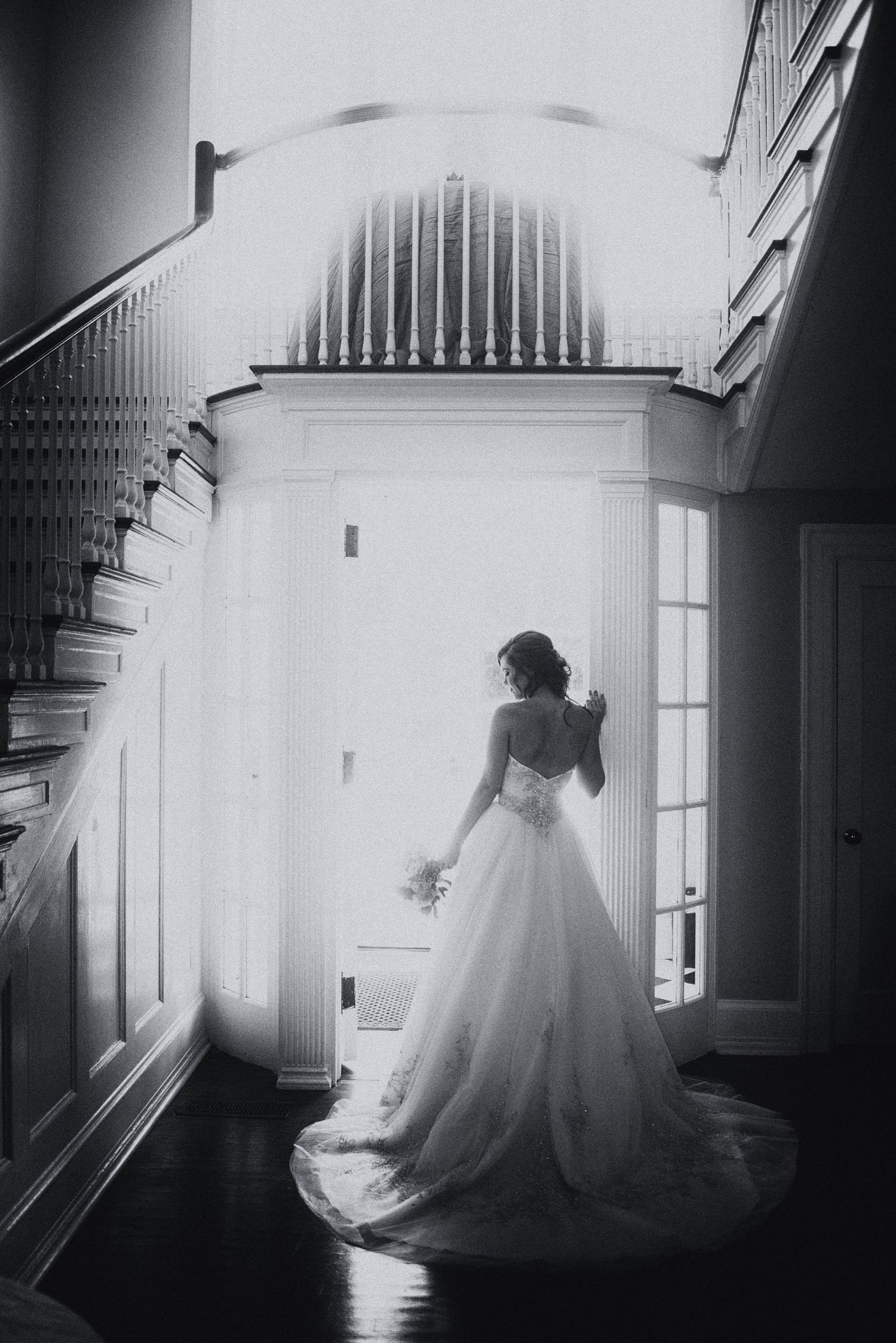 A bride in a flowing white gown stands before a grand doorway in a dramatic black and white wedding portrait.
