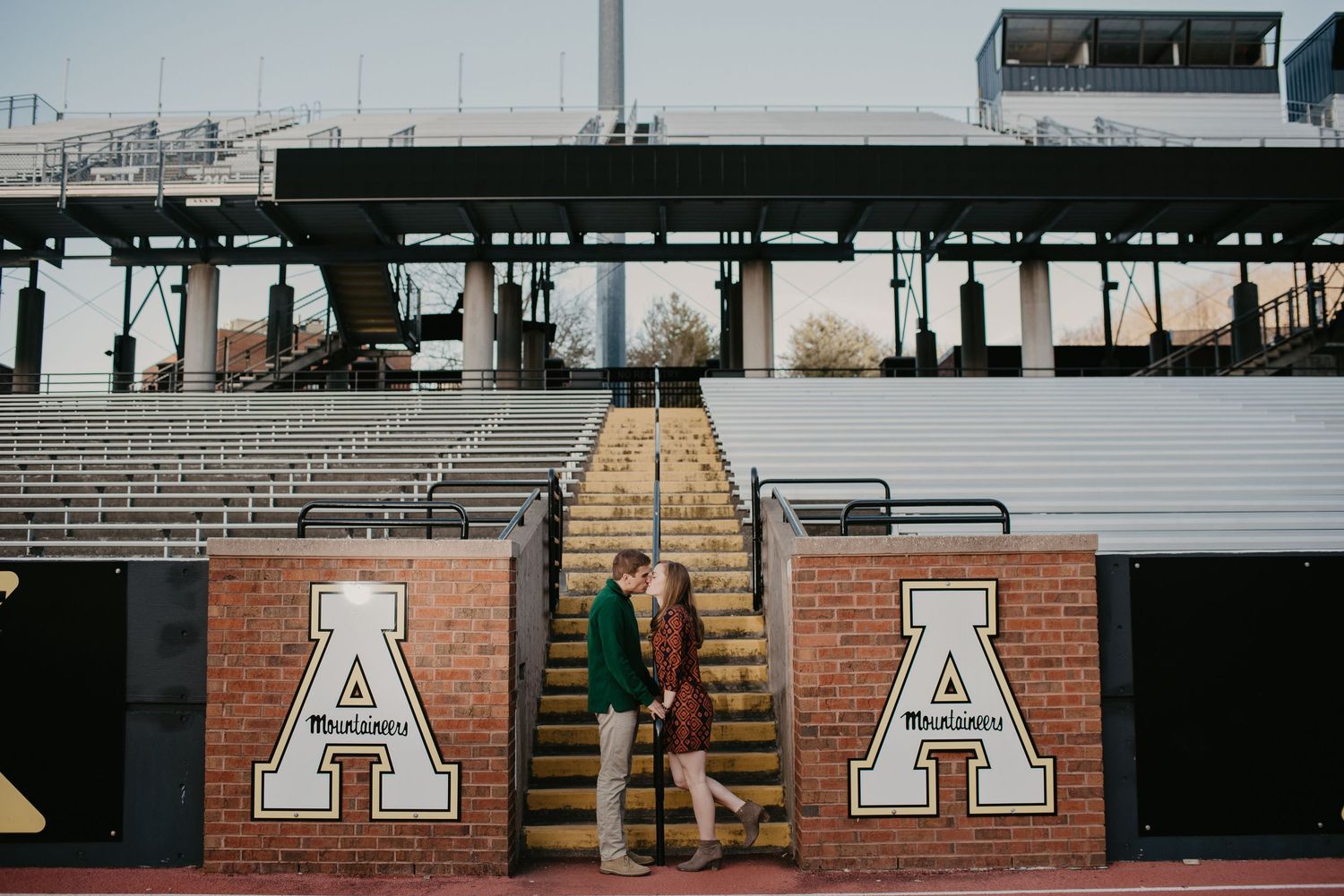 Couple sharing a kiss on stadium stairs beside Appalachian State University brick wall signage.