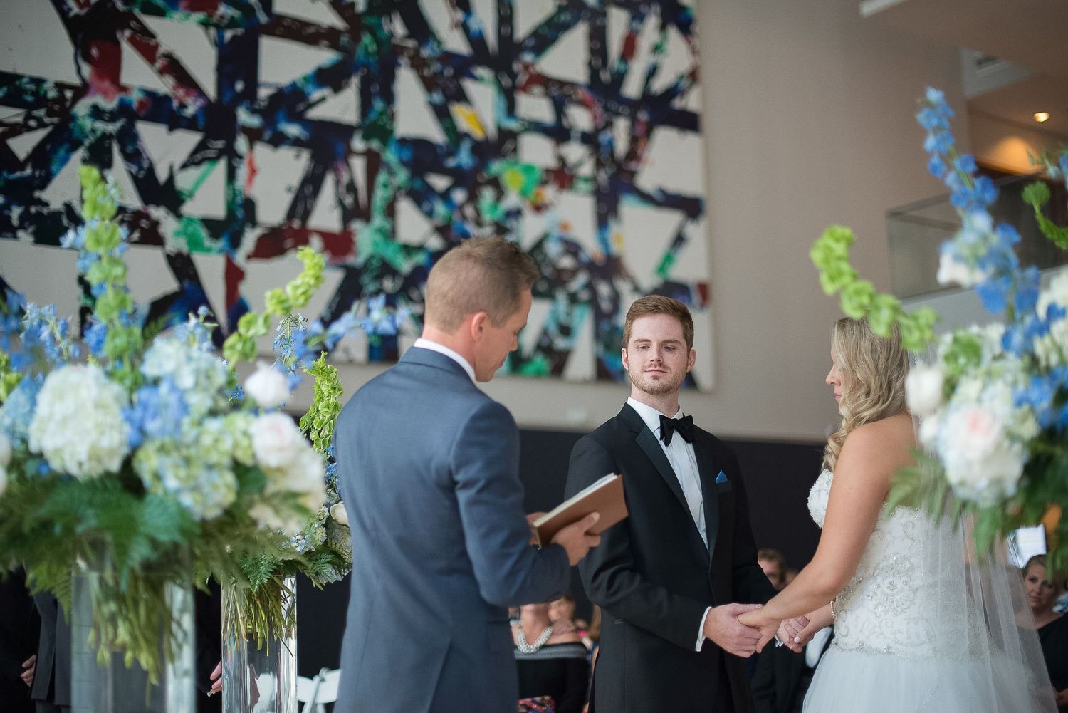 Couple exchanges vows before officiant with blue floral arrangements and colorful artwork backdrop.