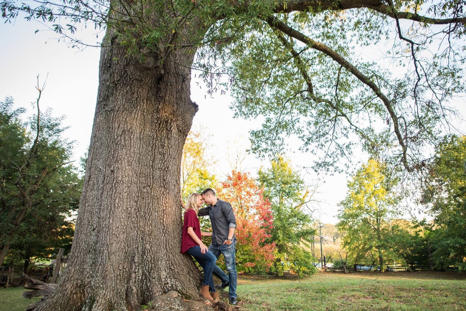 Couple leans against large tree surrounded by colorful autumn foliage.
