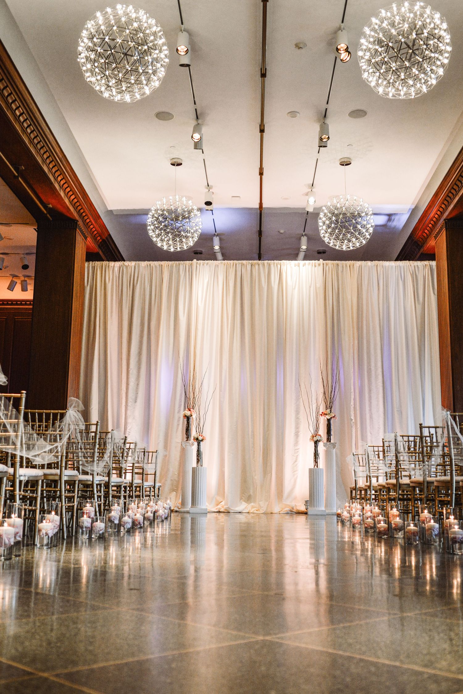 Elegant wedding ceremony venue with white draping backdrop, gold chiavari chairs, and sparkling globe chandeliers.