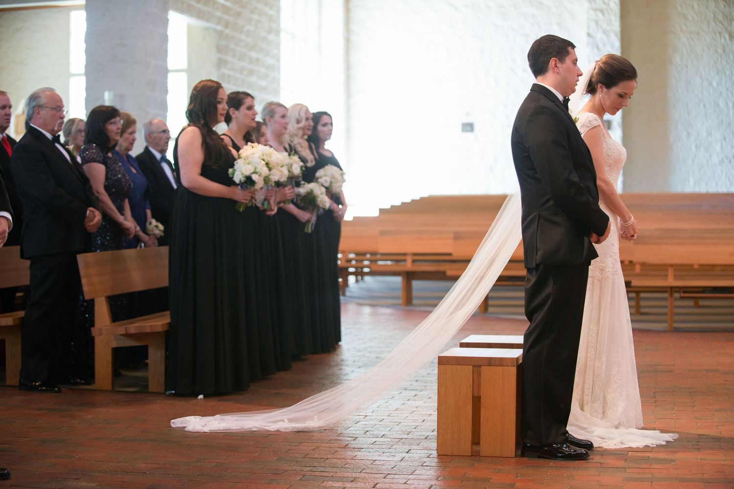 Bride and groom bowing at church altar with bridesmaids in dark gowns during wedding ceremony.