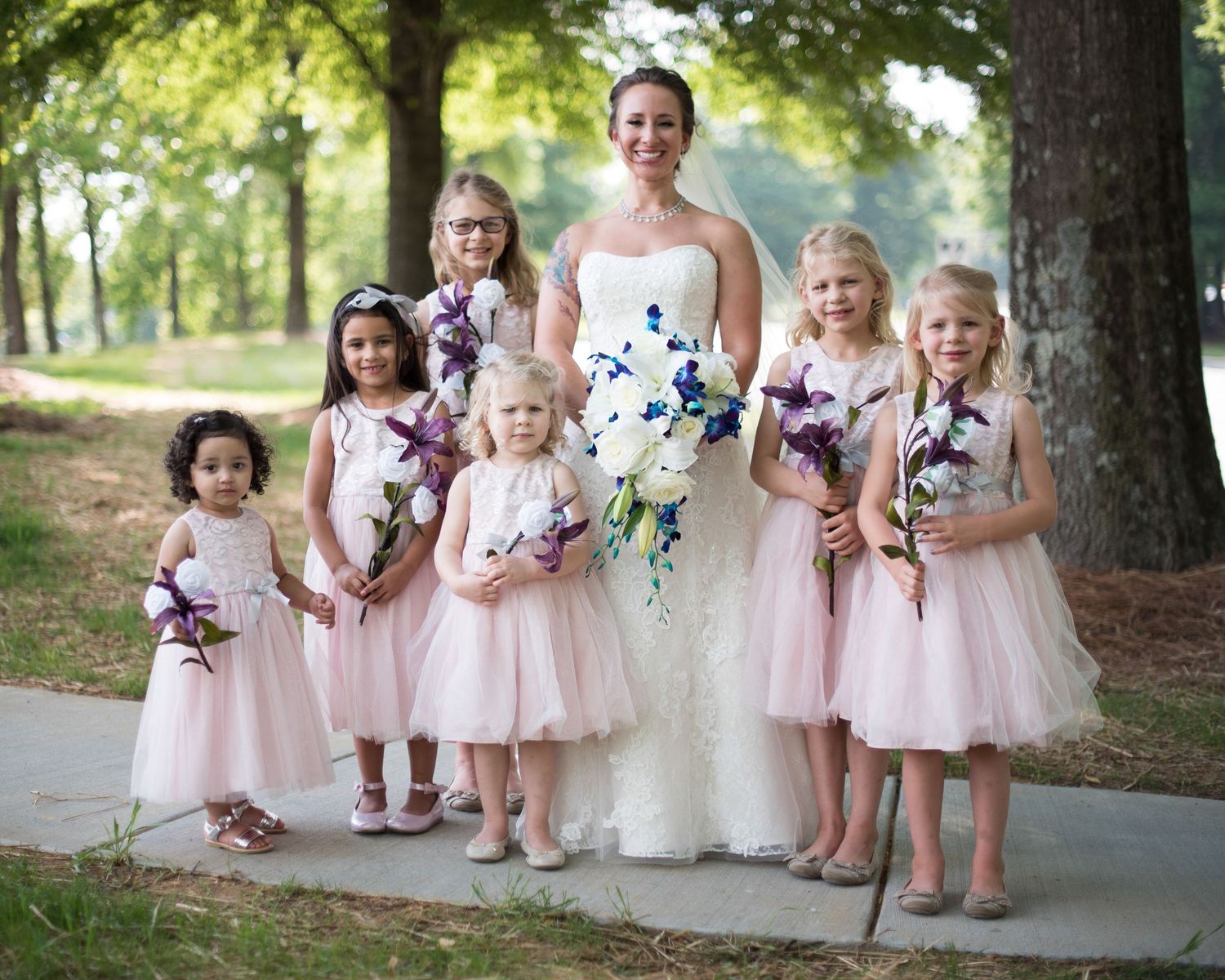 Bride poses with six flower girls in blush pink dresses holding purple and white bouquets outdoors.