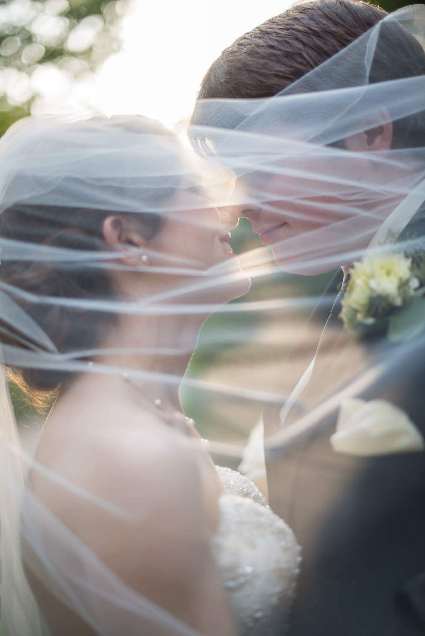 Romantic bride and groom sharing an intimate moment under a flowing wedding veil in golden light.