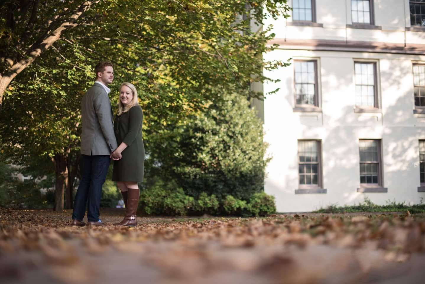 Couple holding hands in autumn leaves outside historic white building with large windows.