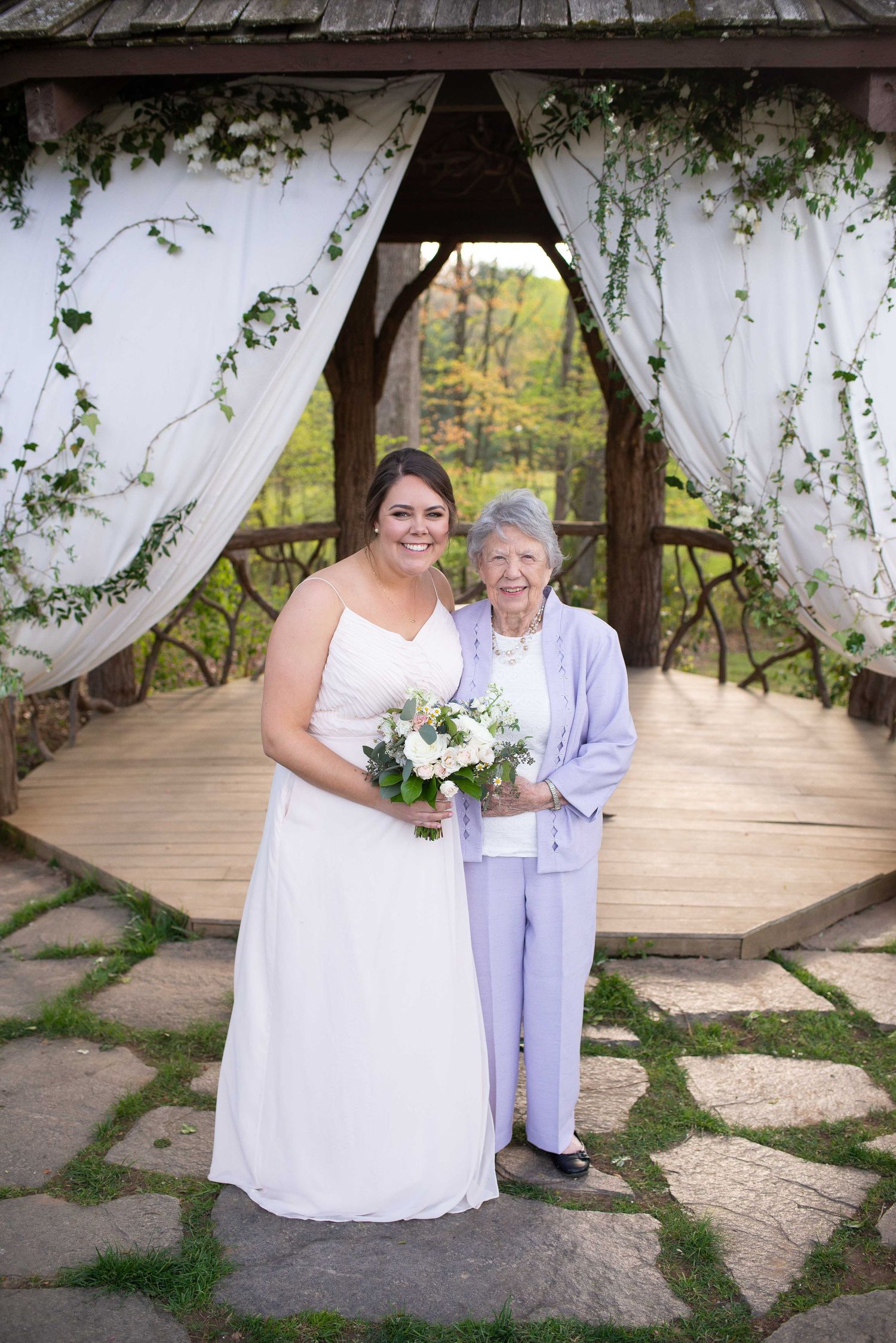 Bride in white gown holding bouquet poses with elderly woman in lavender at decorated outdoor wedding gazebo.