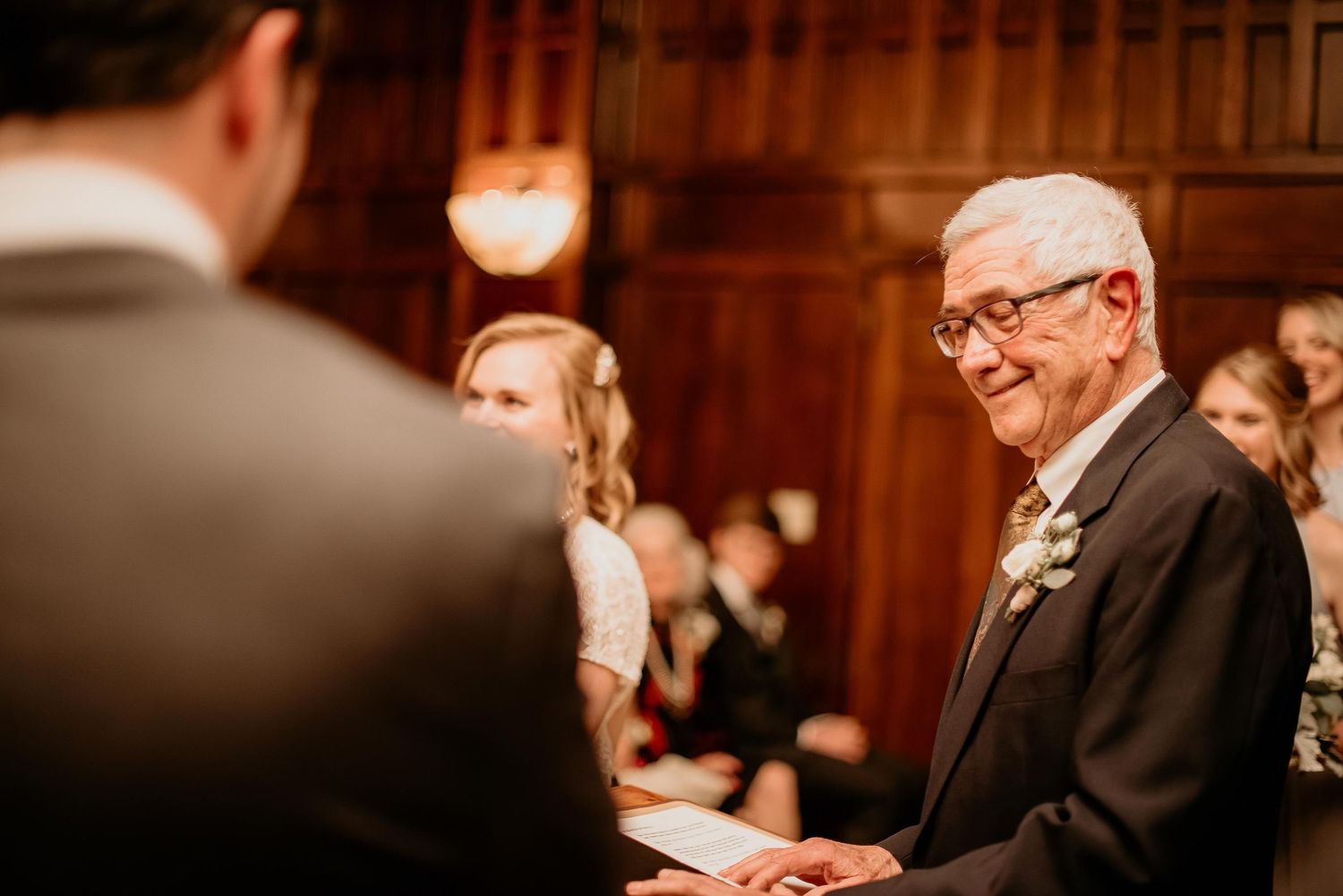Elderly man smiling warmly during a wedding ceremony in an elegant wood-paneled venue.