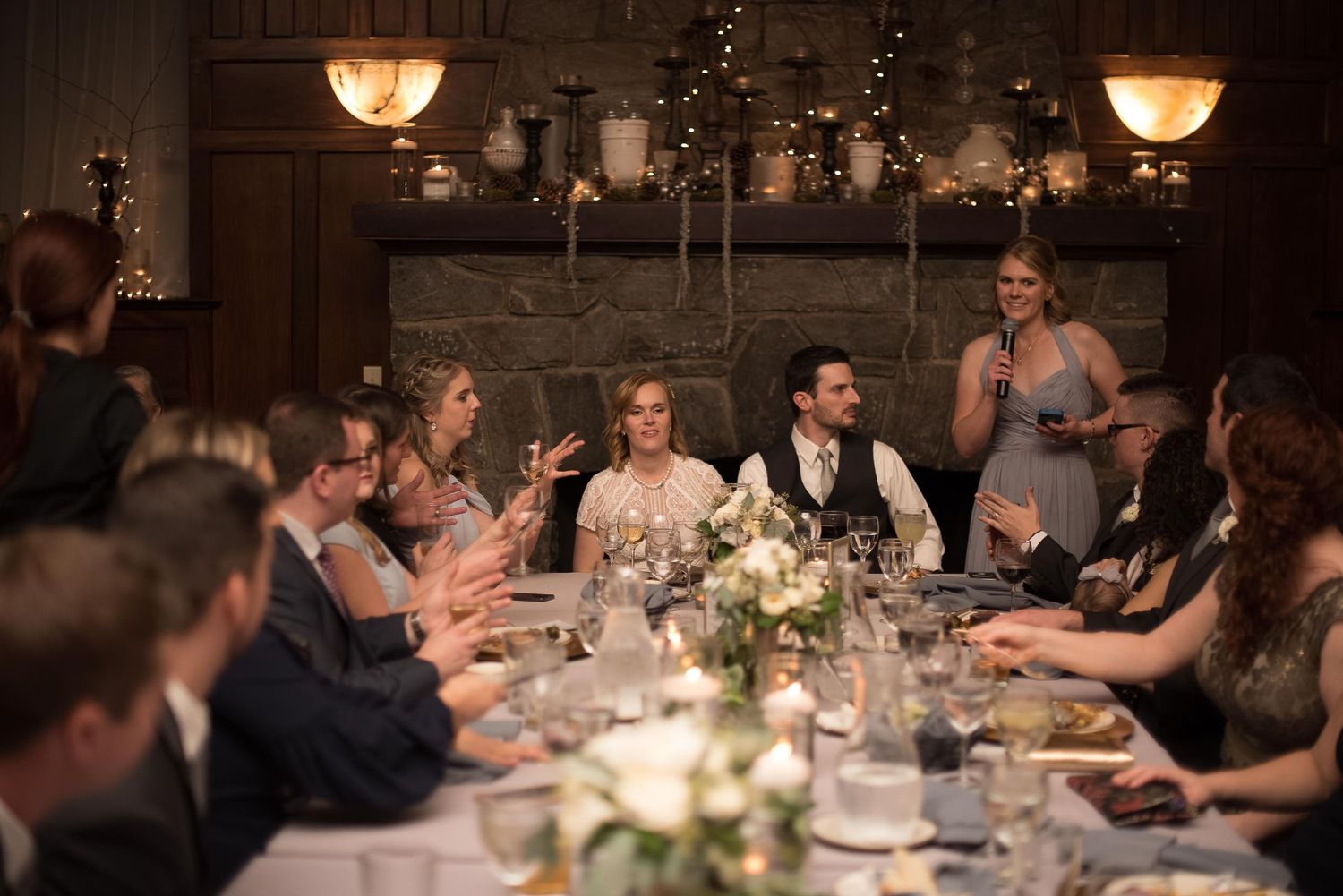 Wedding reception dinner party with guests toasting at an elegant candlelit table near a stone fireplace.