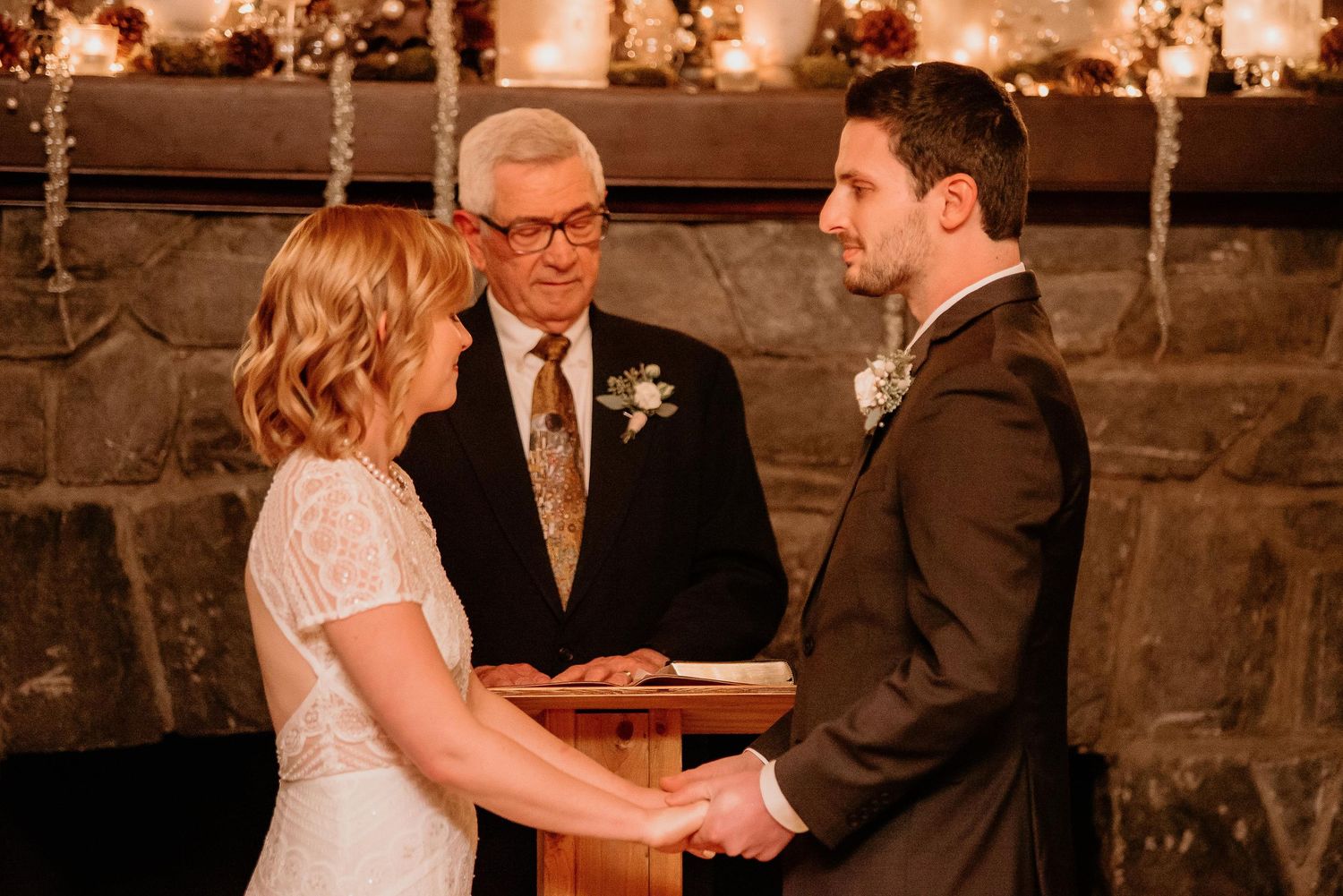 Bride and groom exchange vows before officiant at rustic stone fireplace wedding ceremony.