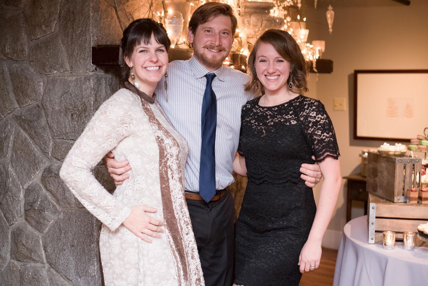Three smiling friends pose together at an elegant indoor event, with two women in dresses flanking a man in a tie.