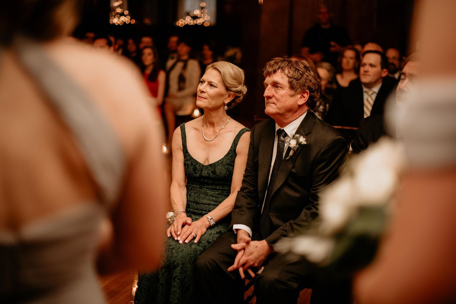 Elegant wedding guests seated in a warmly lit ceremony venue, an older couple in formal attire watching the proceedings.