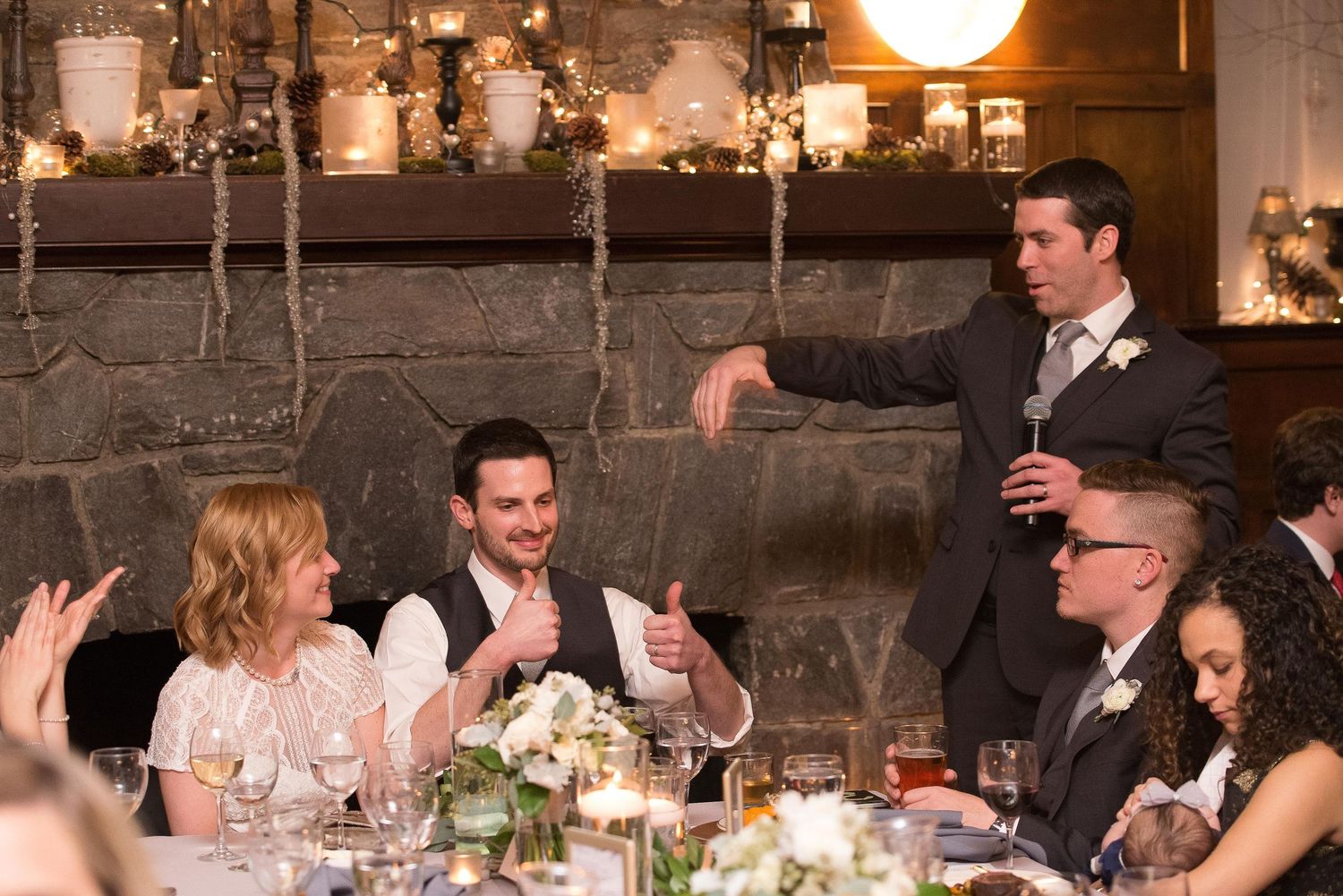 Man giving a toast at an elegant wedding reception dinner with guests laughing and applauding by a stone fireplace.