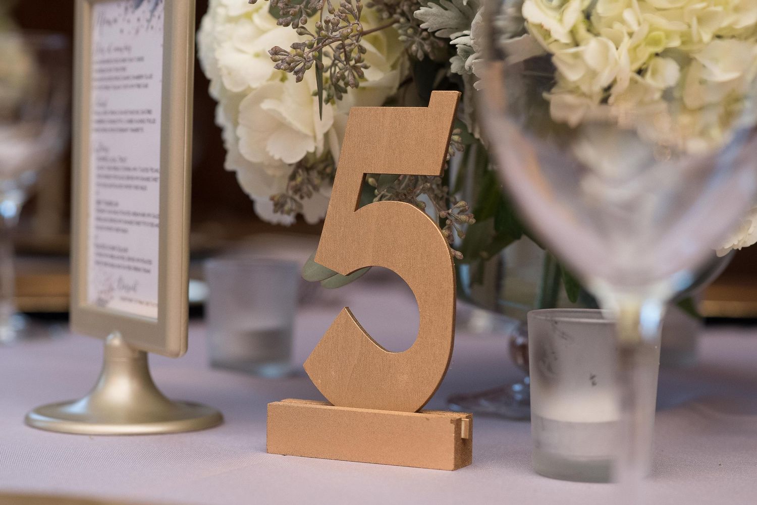 Gold number 5 table marker at elegant wedding reception with white floral centerpiece and glassware in background.