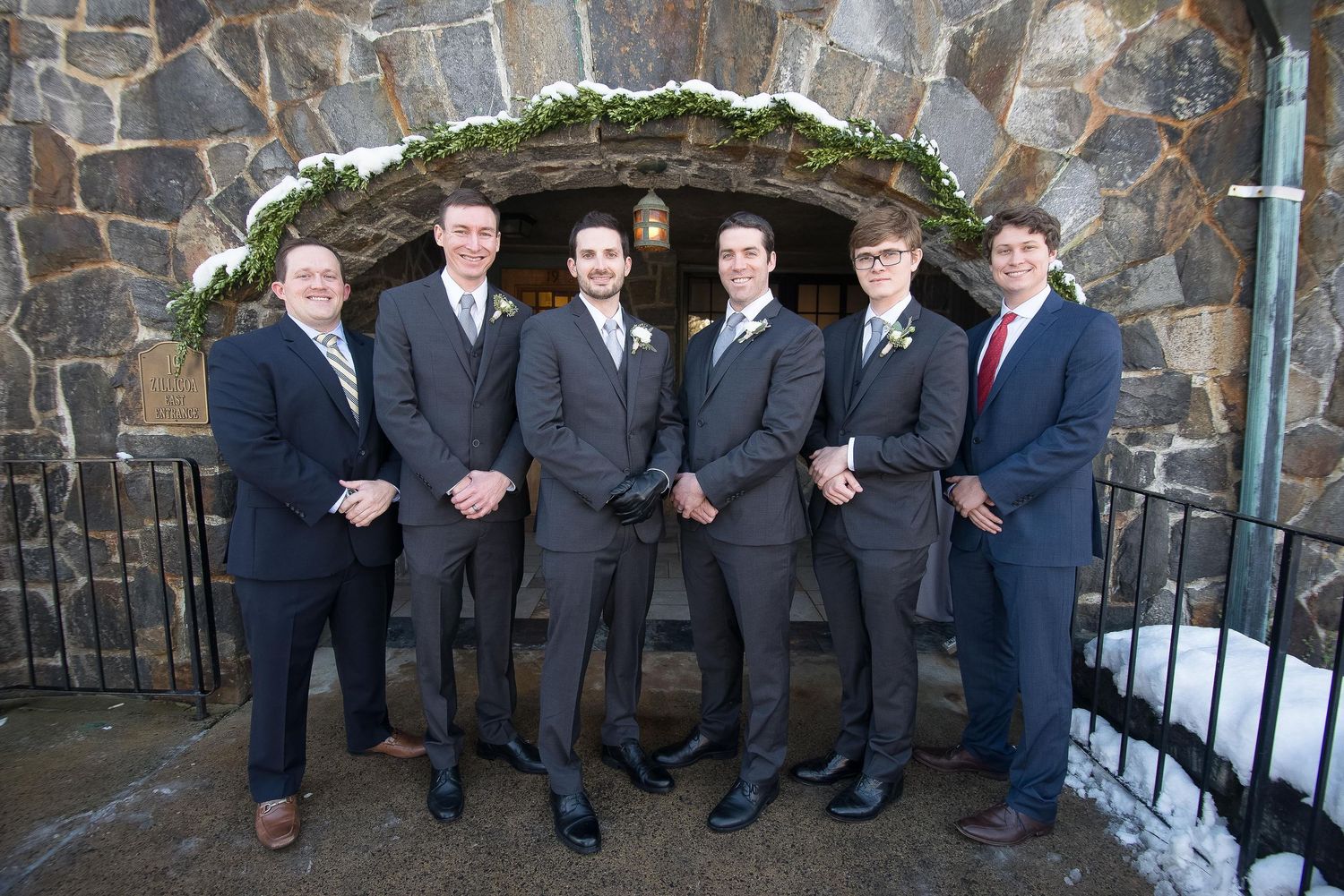 Five groomsmen in gray suits posing together in front of a stone archway at a winter wedding venue.
