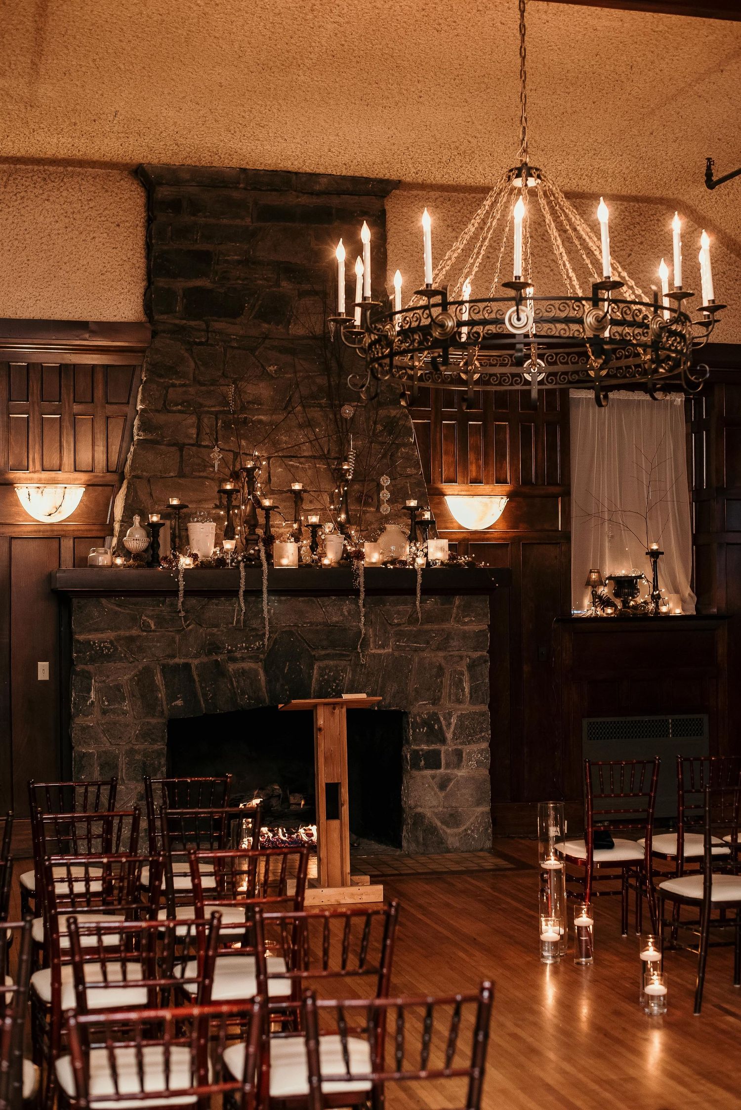 Elegant indoor wedding ceremony with stone fireplace, candlelit chandelier, and dark wood Chiavari chairs.