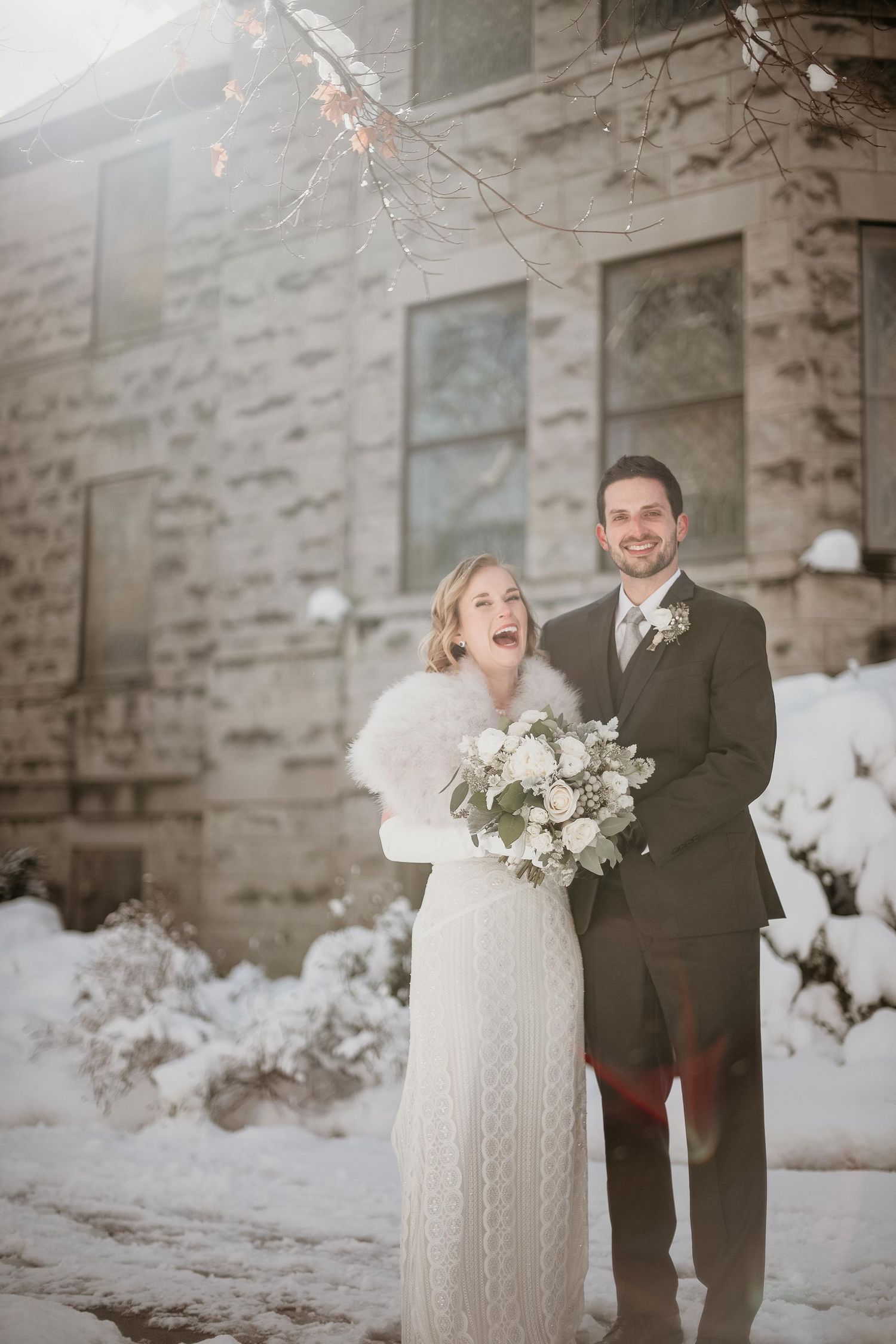 Joyful bride and groom pose in snow outside stone building, bride in white lace gown with fur wrap holding bouquet.
