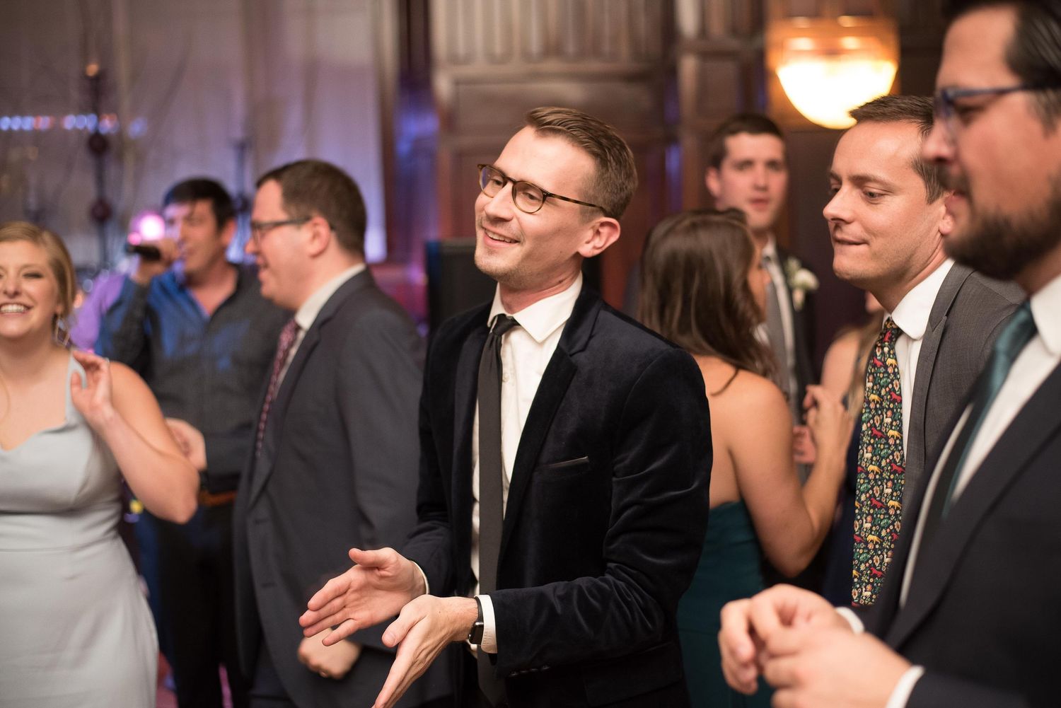 Man in black suit smiling and clapping at a lively wedding reception dance floor with guests in background.