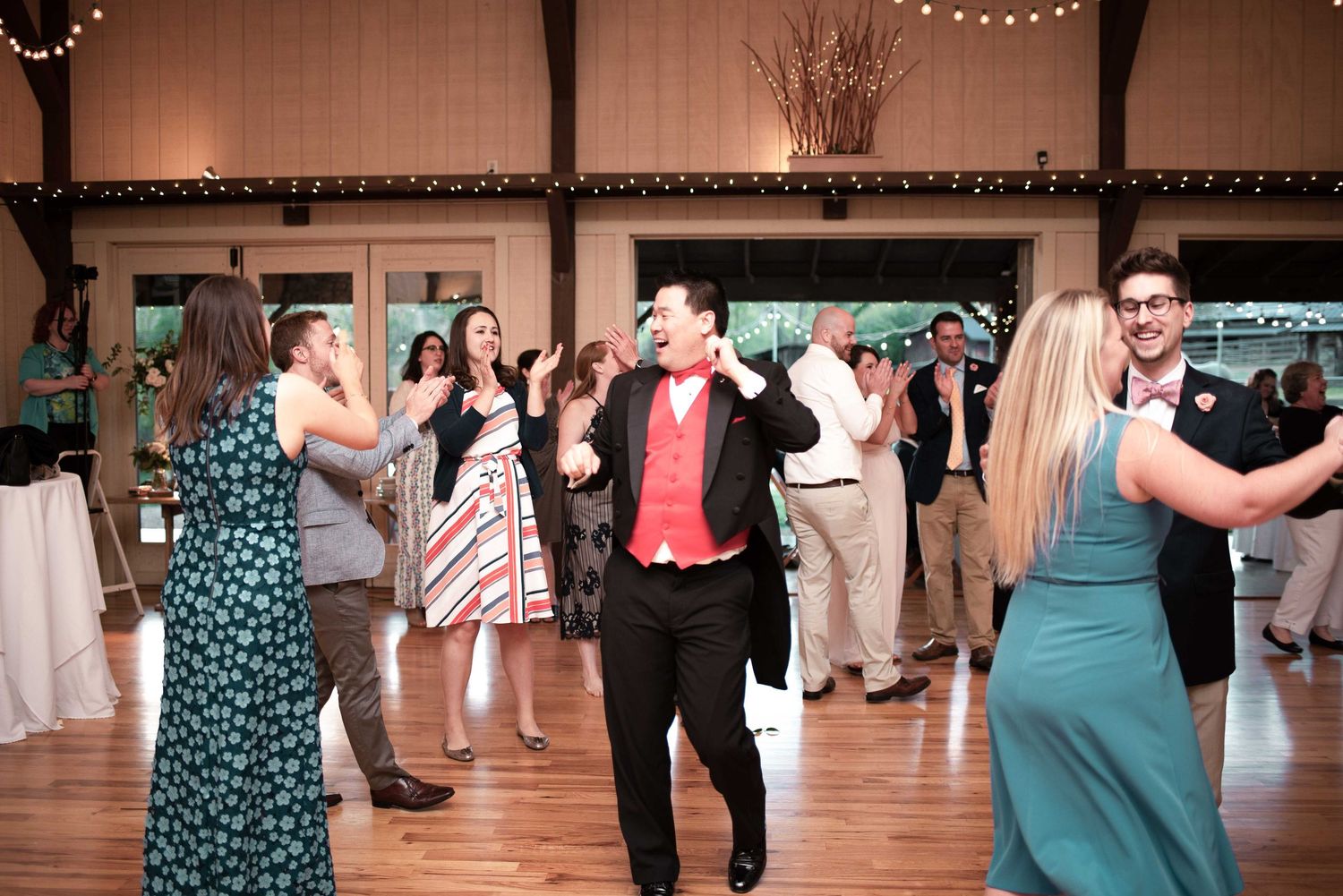 Guests dancing joyfully at a wedding reception in a rustic barn venue with string lights.