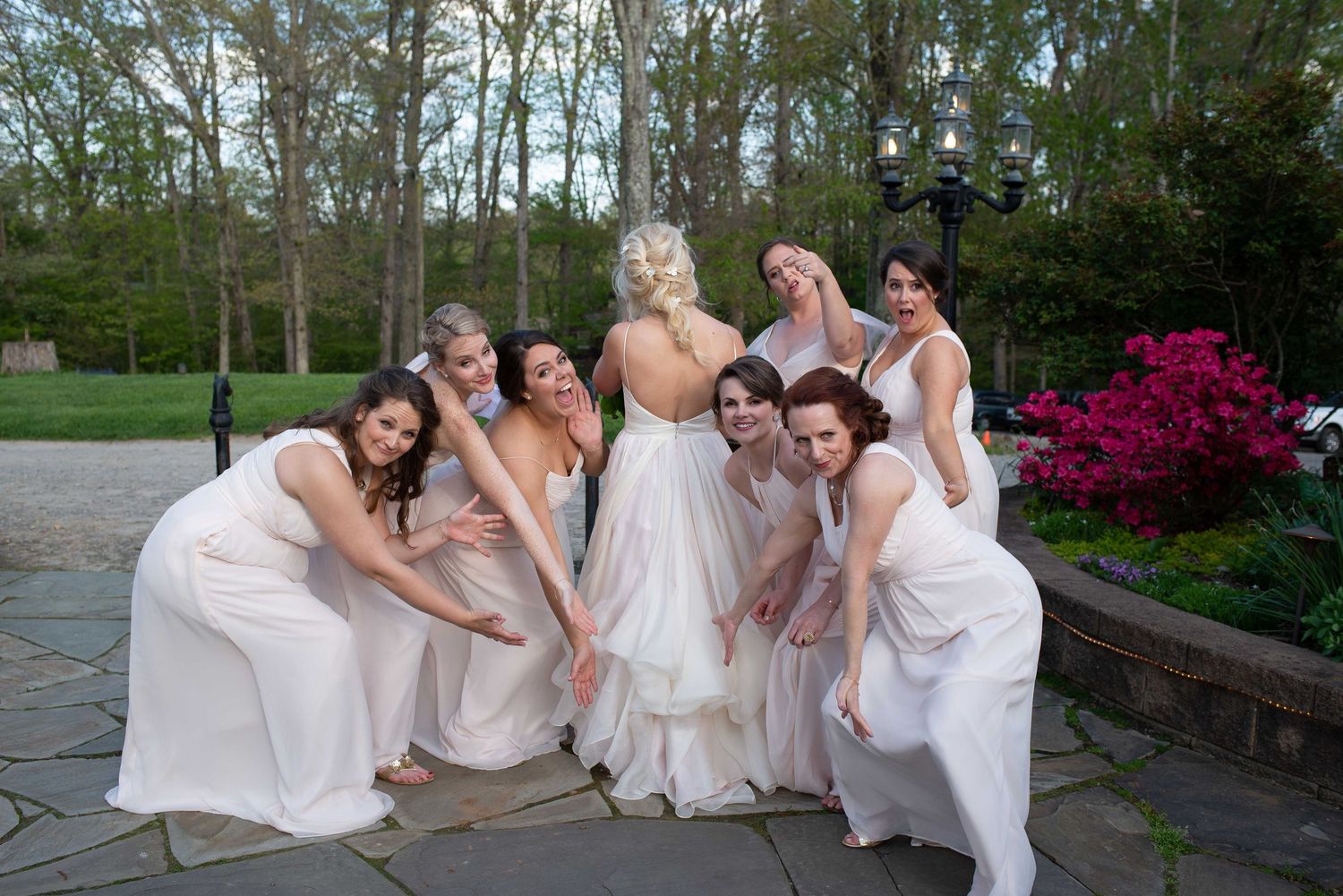 Bride in white gown poses with smiling bridesmaids in white dresses outdoors at a garden wedding venue.