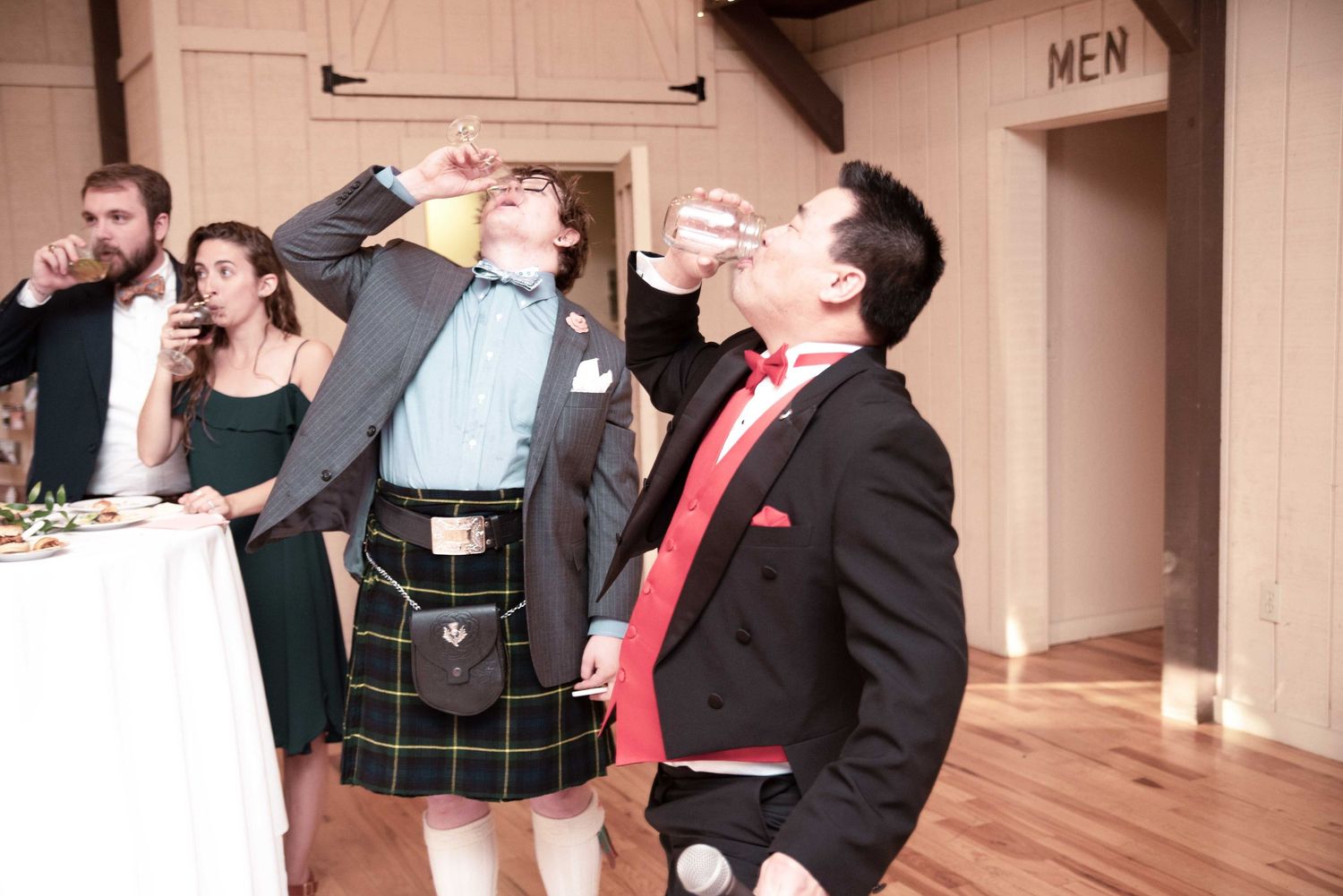 Two men doing shots at a wedding reception, one wearing a kilt and one in a tuxedo with red tie.