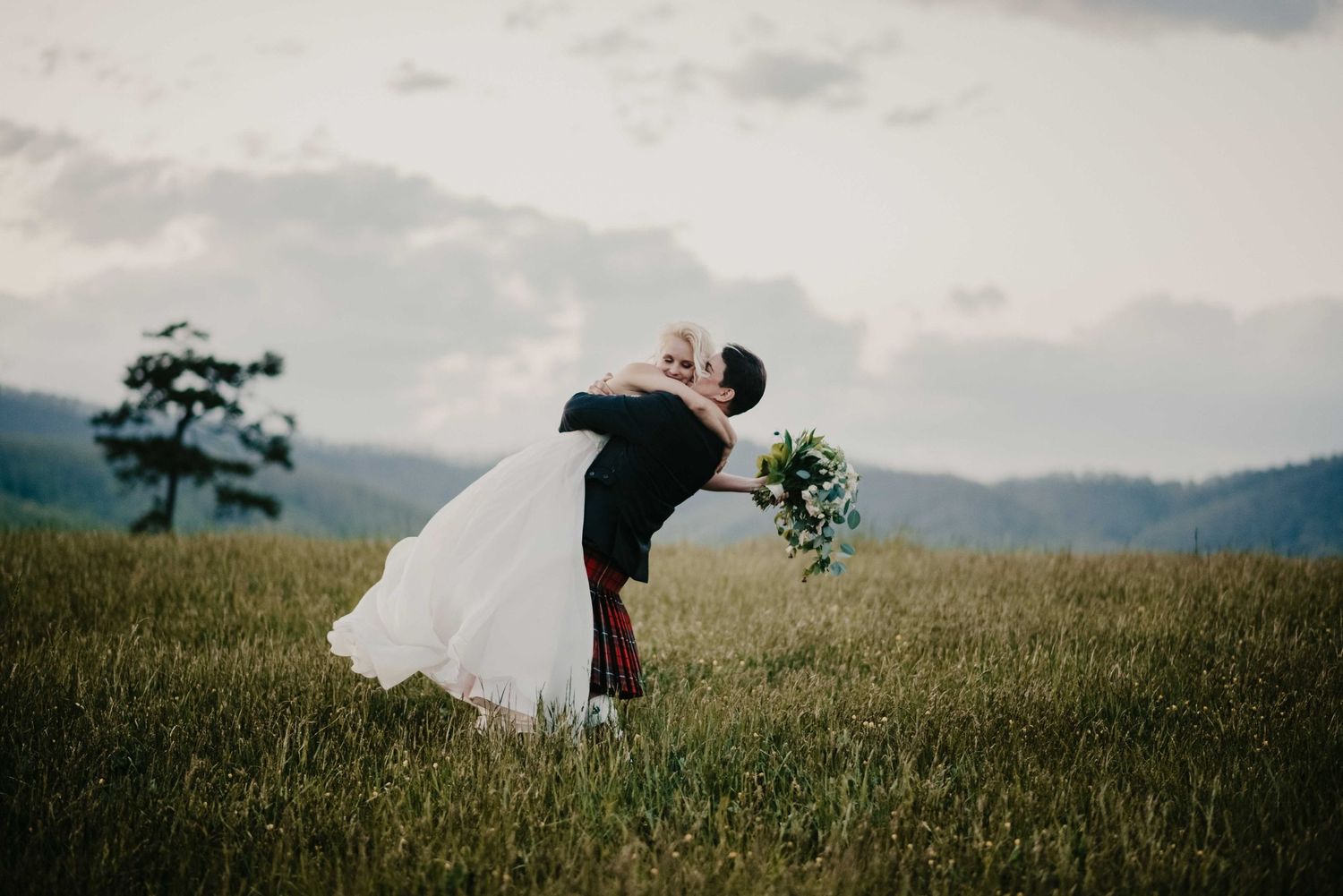 Groom dipping bride for a kiss in a lush green mountain meadow on their wedding day.