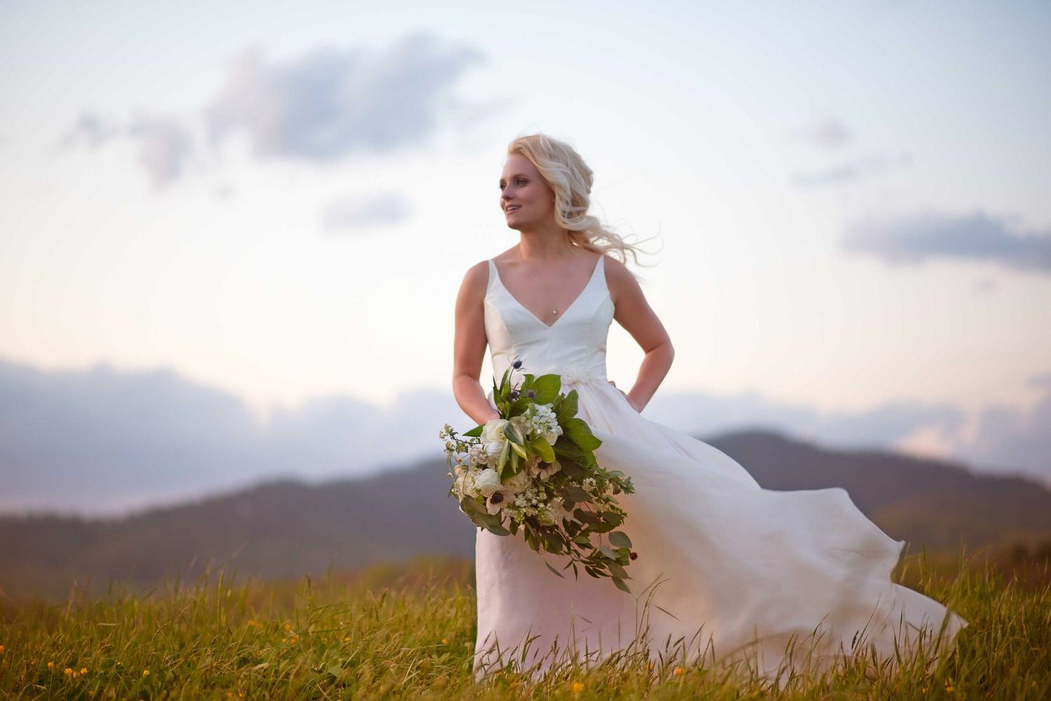 Bride in white gown holding bouquet stands in mountain meadow at sunset with wind blowing her dress.