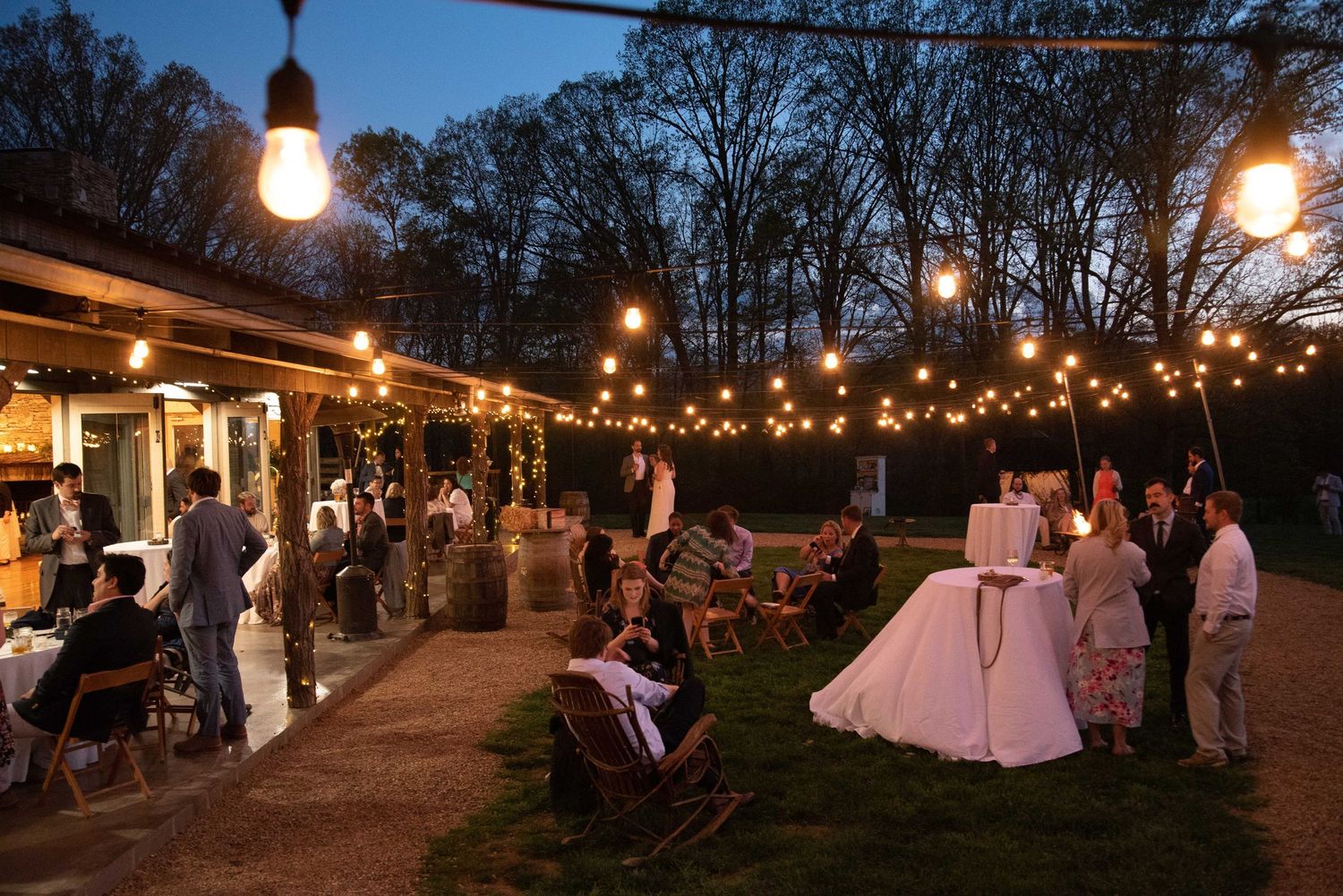 Outdoor evening wedding reception with string lights, guests mingling, and a bride in white dress in a garden setting.