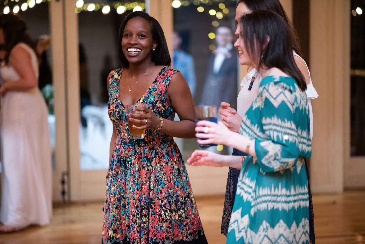 Two women laughing and holding drinks at a festive indoor party, wearing colorful dresses in a warmly lit venue.