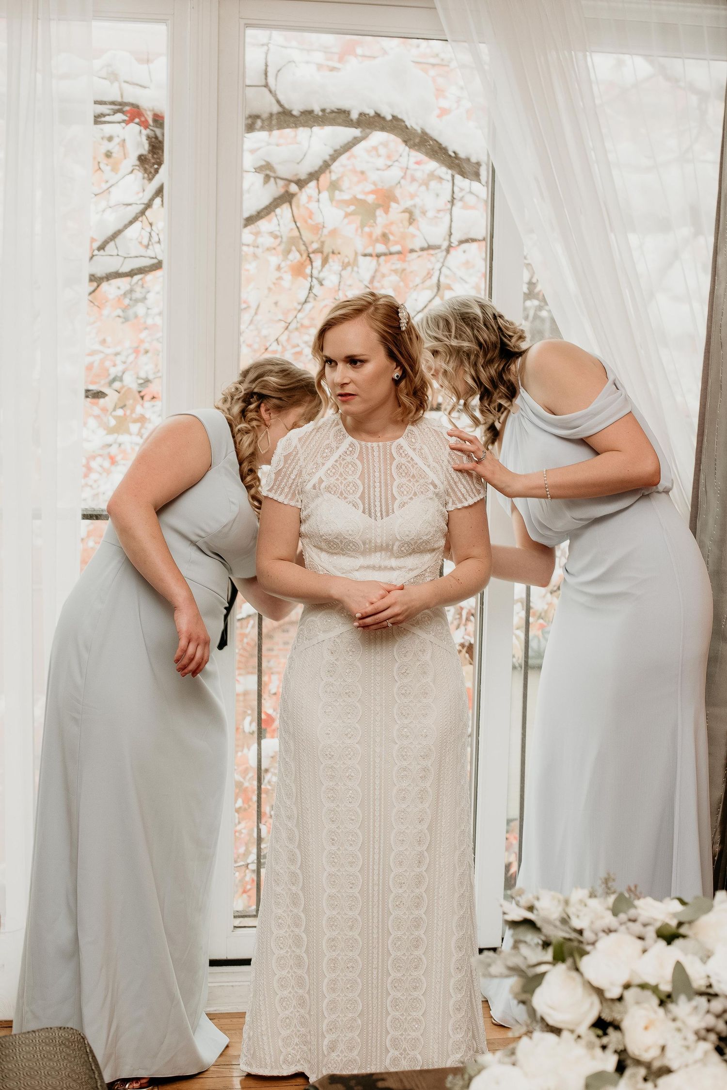 Bride in white lace gown being assisted by two bridesmaids in silver dresses near a window with floral backdrop.