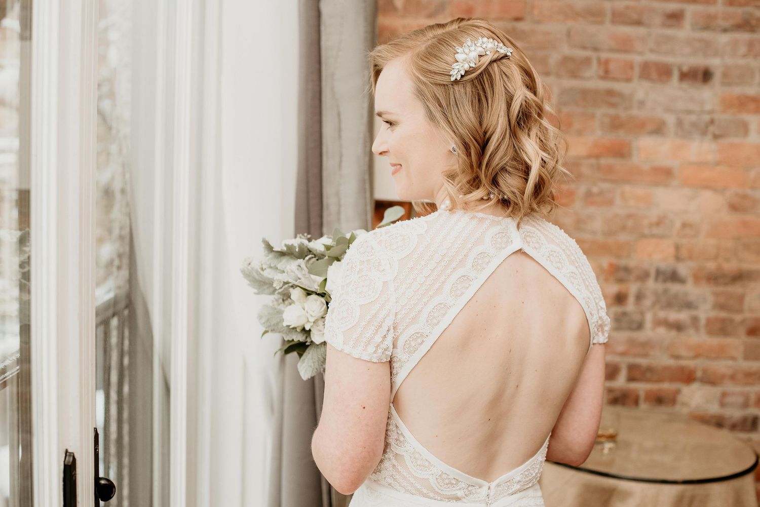 Bride in backless beaded wedding gown holding white bouquet, wearing tiara, standing by window with brick wall backdrop.