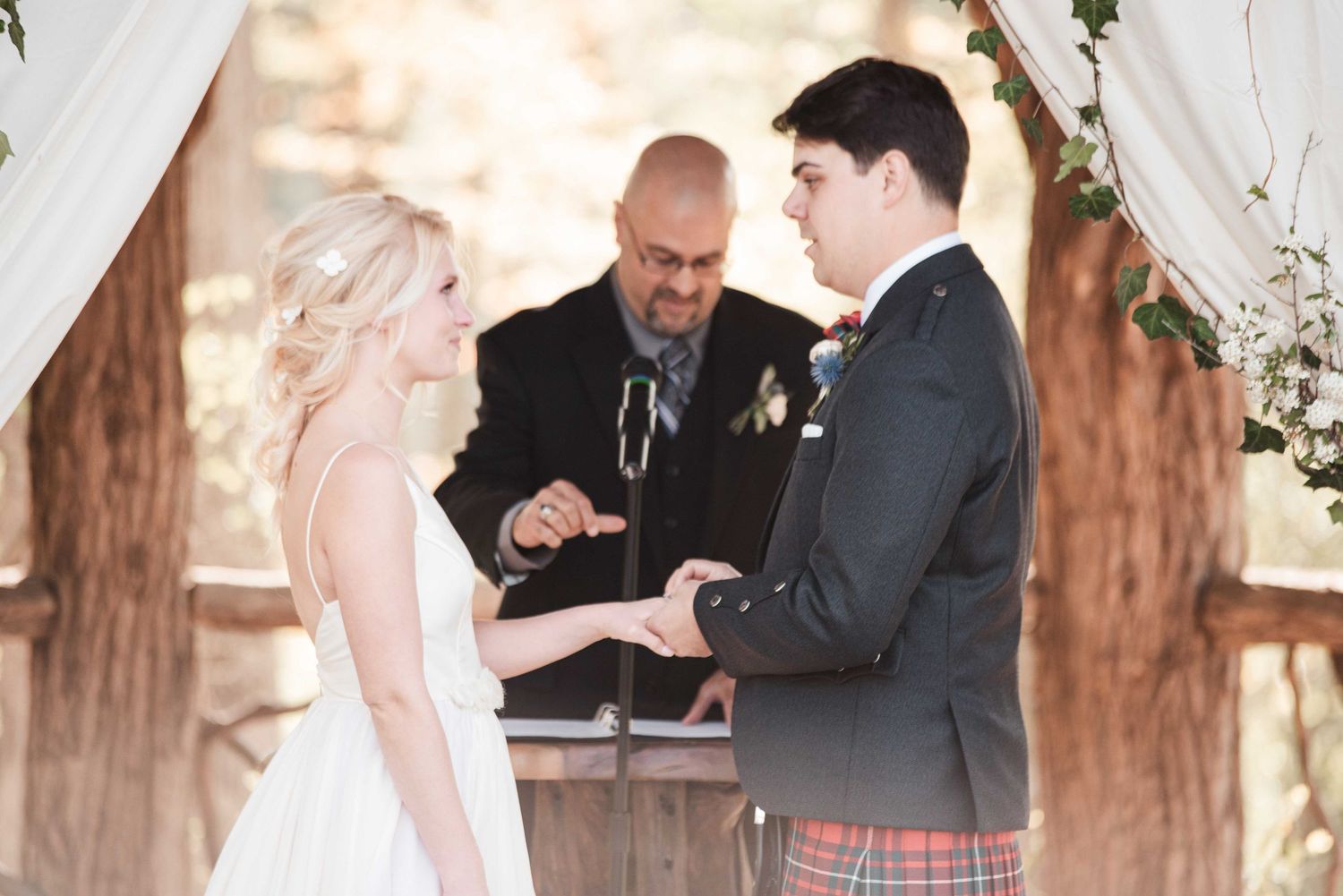 Groom in tartan kilt exchanges rings with bride at outdoor wedding ceremony officiated by minister.