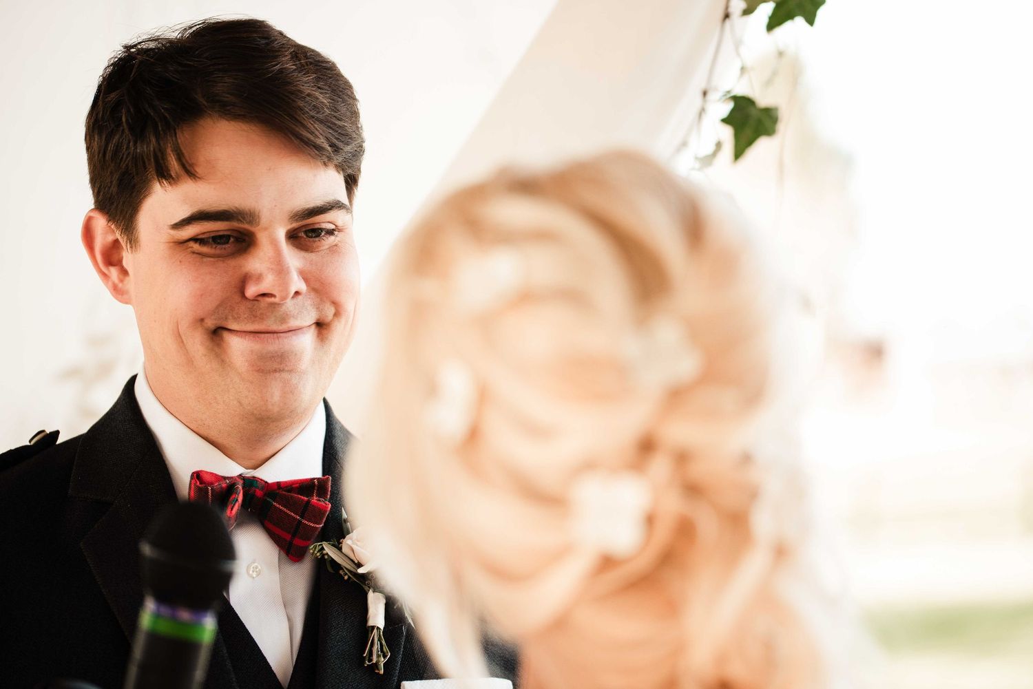Groom in black suit and red tartan bow tie smiling at blonde bride during wedding ceremony.