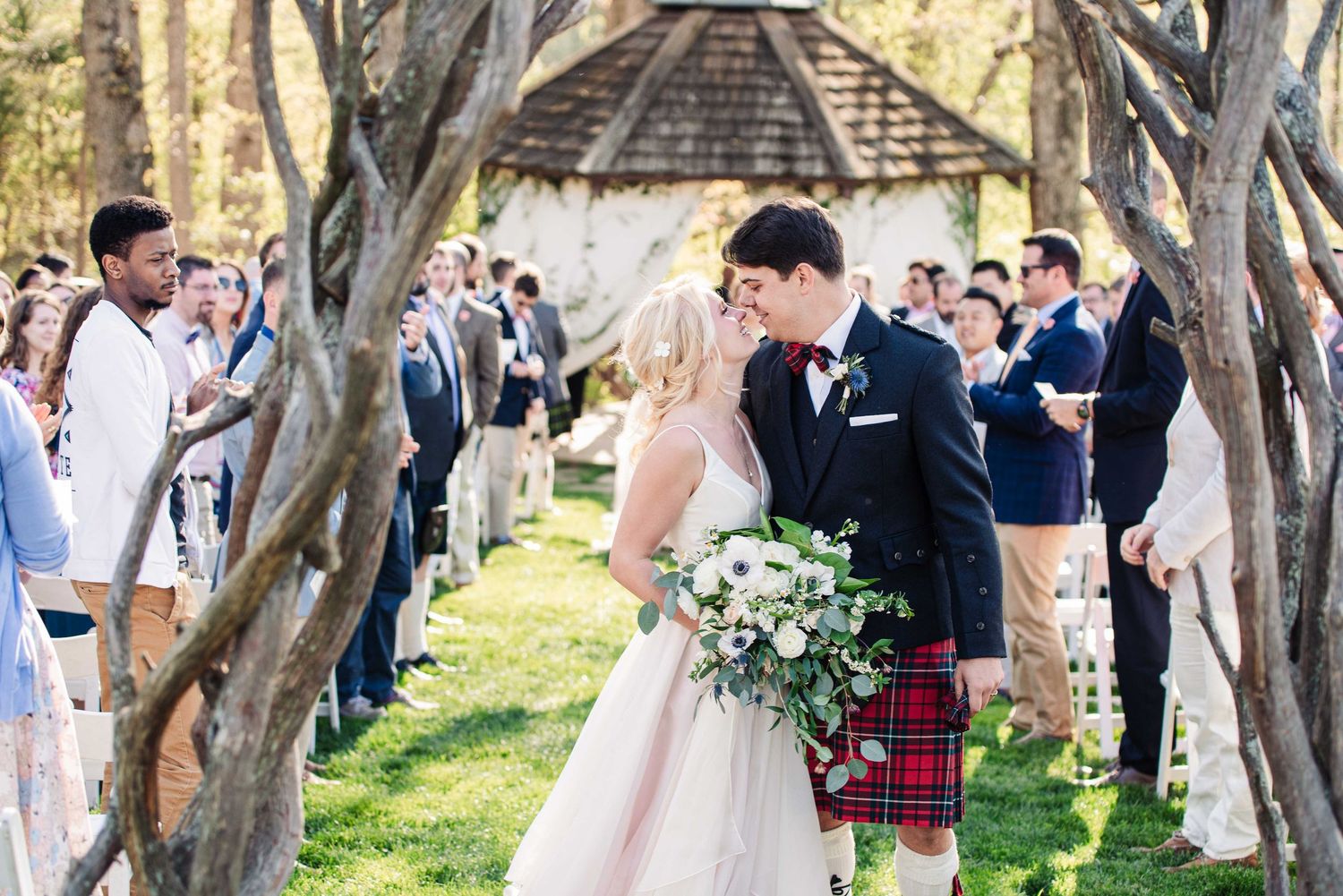 Bride and groom share a kiss at outdoor wedding ceremony, groom wearing traditional Scottish tartan kilt, surrounded by guests.