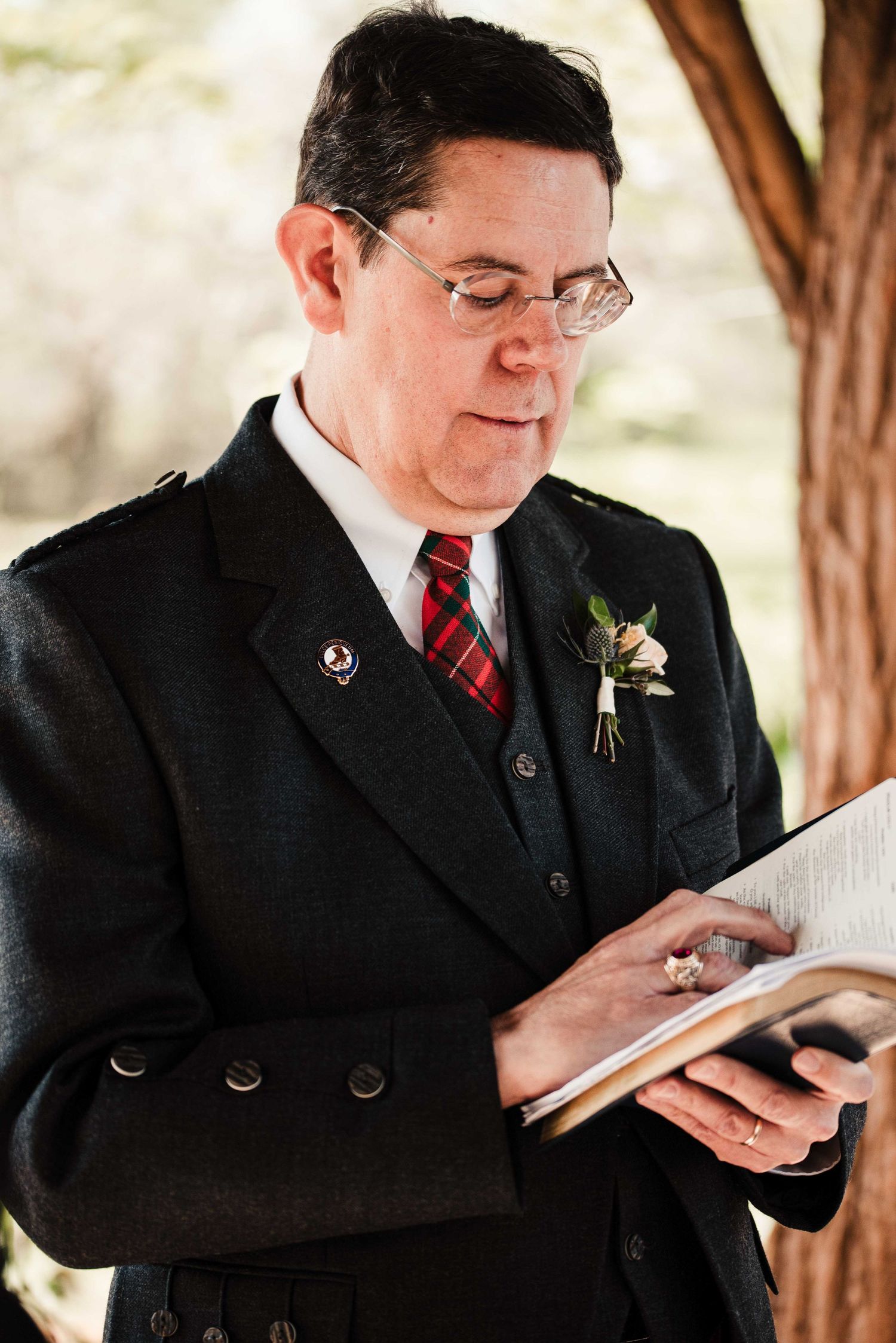 Officiant in dark suit with tartan tie and boutonniere reads from book during outdoor wedding ceremony.