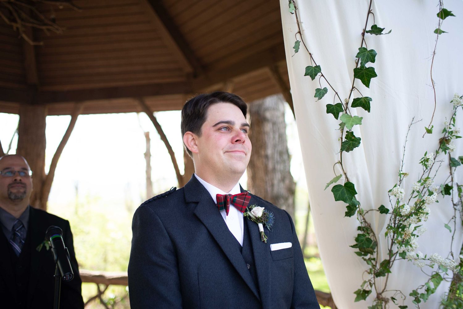 Groom in black suit and burgundy bow tie smiling at wedding ceremony under decorated gazebo with ivy.