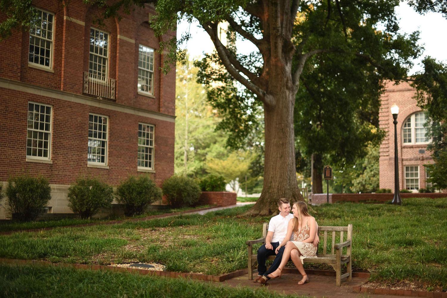 Couple sitting together on a bench beneath a large tree on a scenic brick university campus.