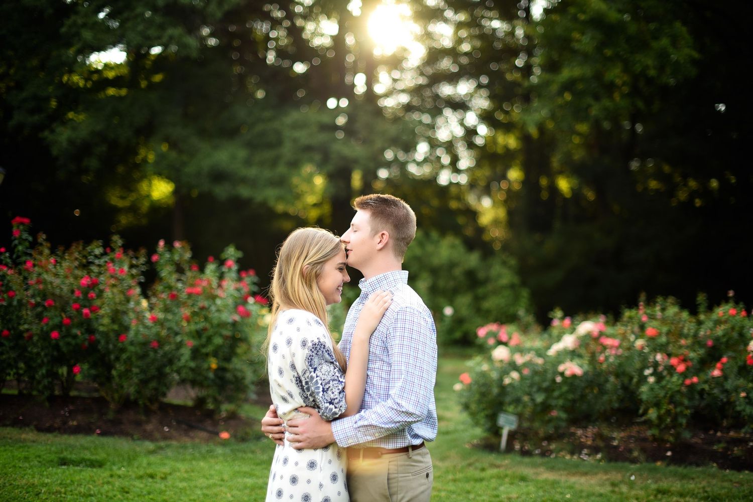 Couple sharing a romantic moment in a lush rose garden with golden sunlight filtering through trees.