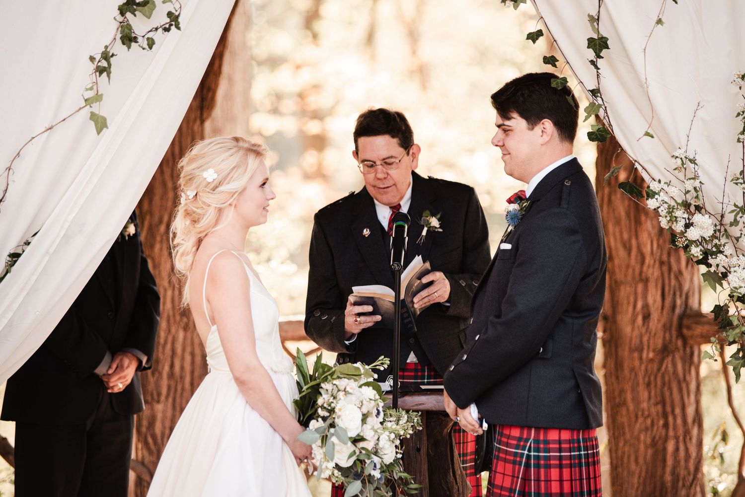 Bride and groom exchange vows at outdoor woodland wedding ceremony with officiant in kilt.