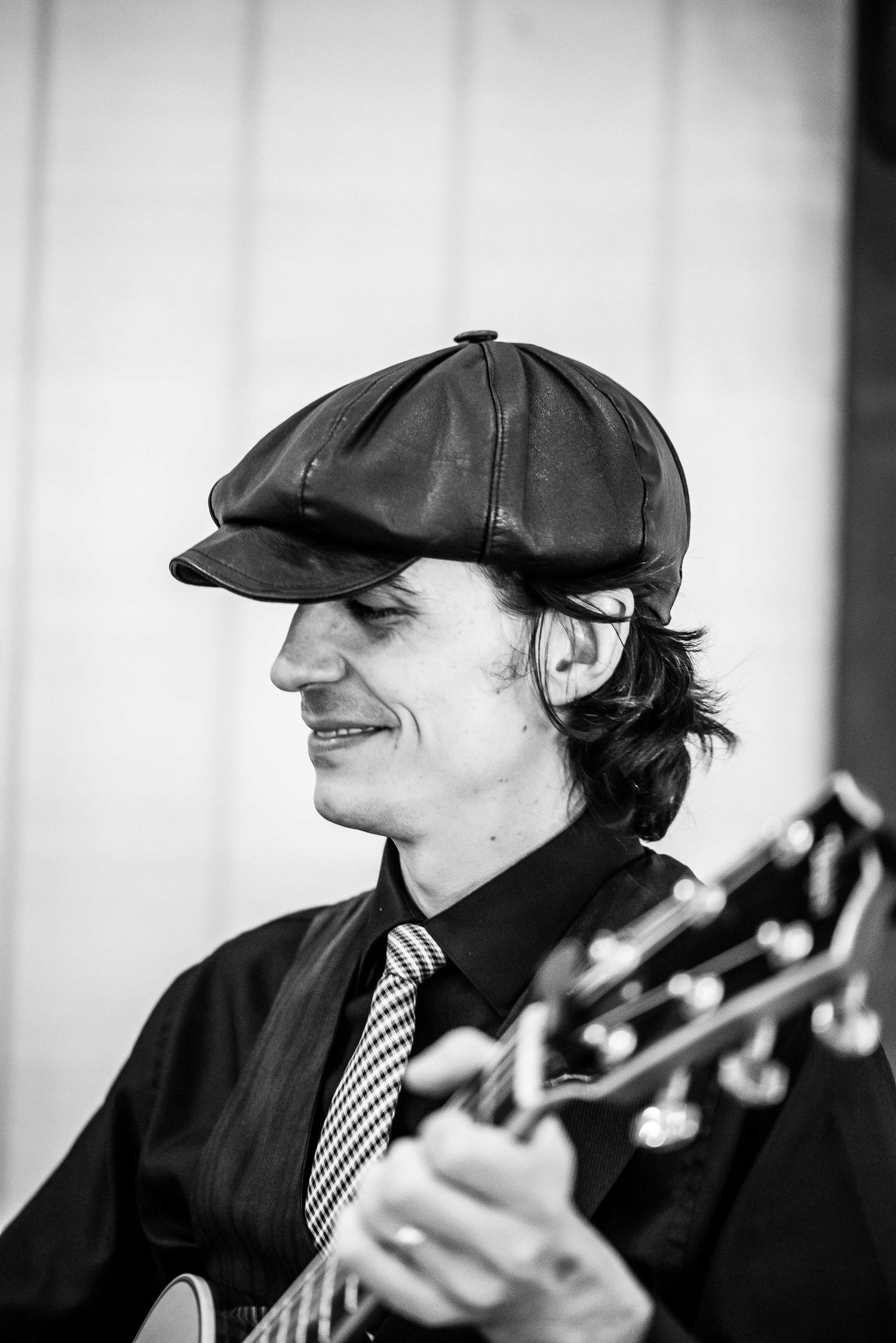 Black and white portrait of a smiling male guitarist wearing a leather flat cap and striped tie at a wedding.