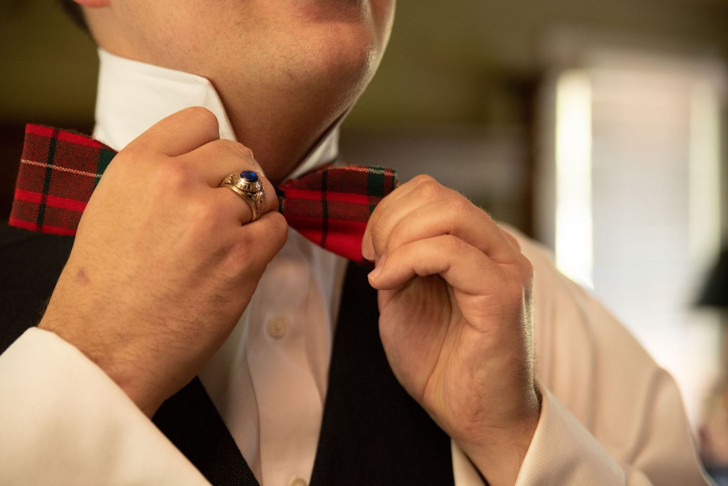 Groom adjusts red plaid bow tie while wearing black vest and white dress shirt.