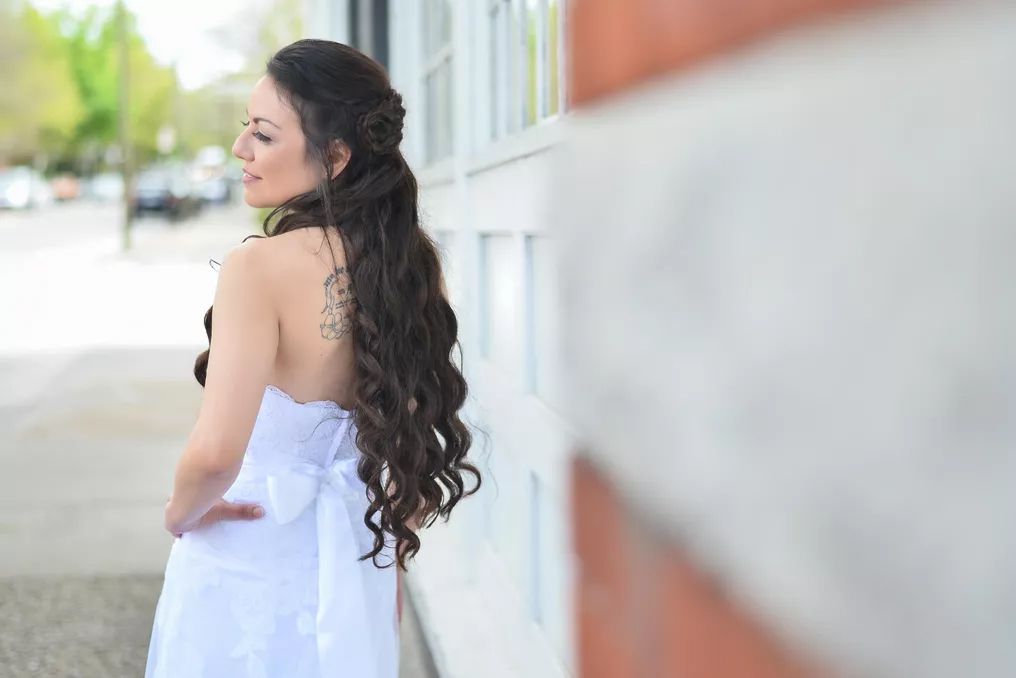 Bride in strapless white gown with back tattoo leaning against brick wall outdoors.