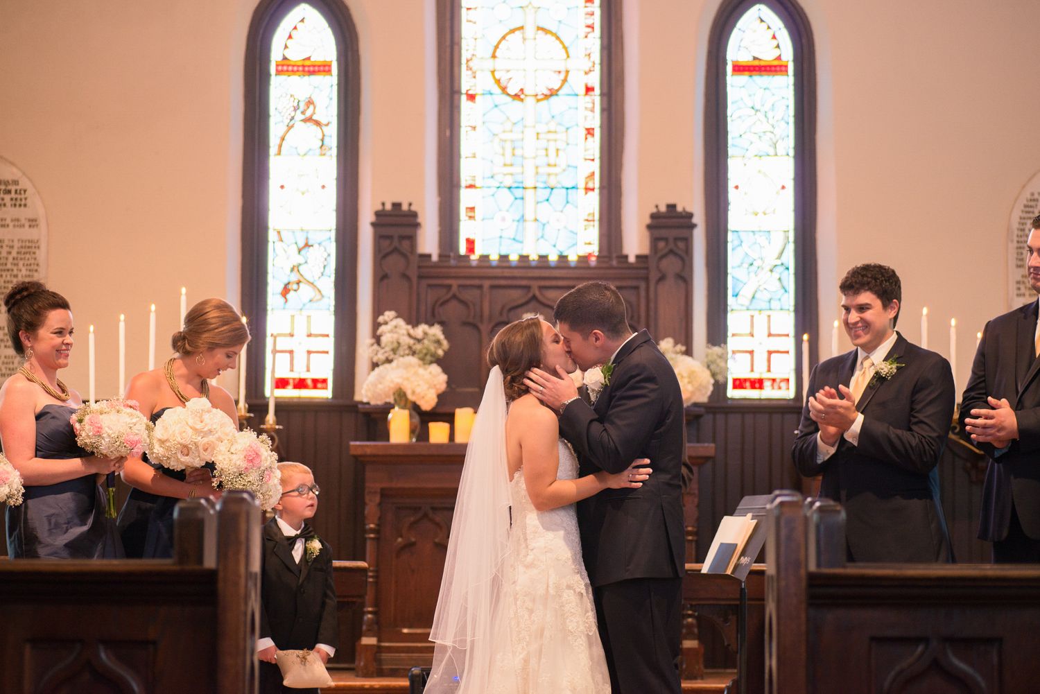Bride and groom share first kiss at church altar during wedding ceremony with stained glass windows.