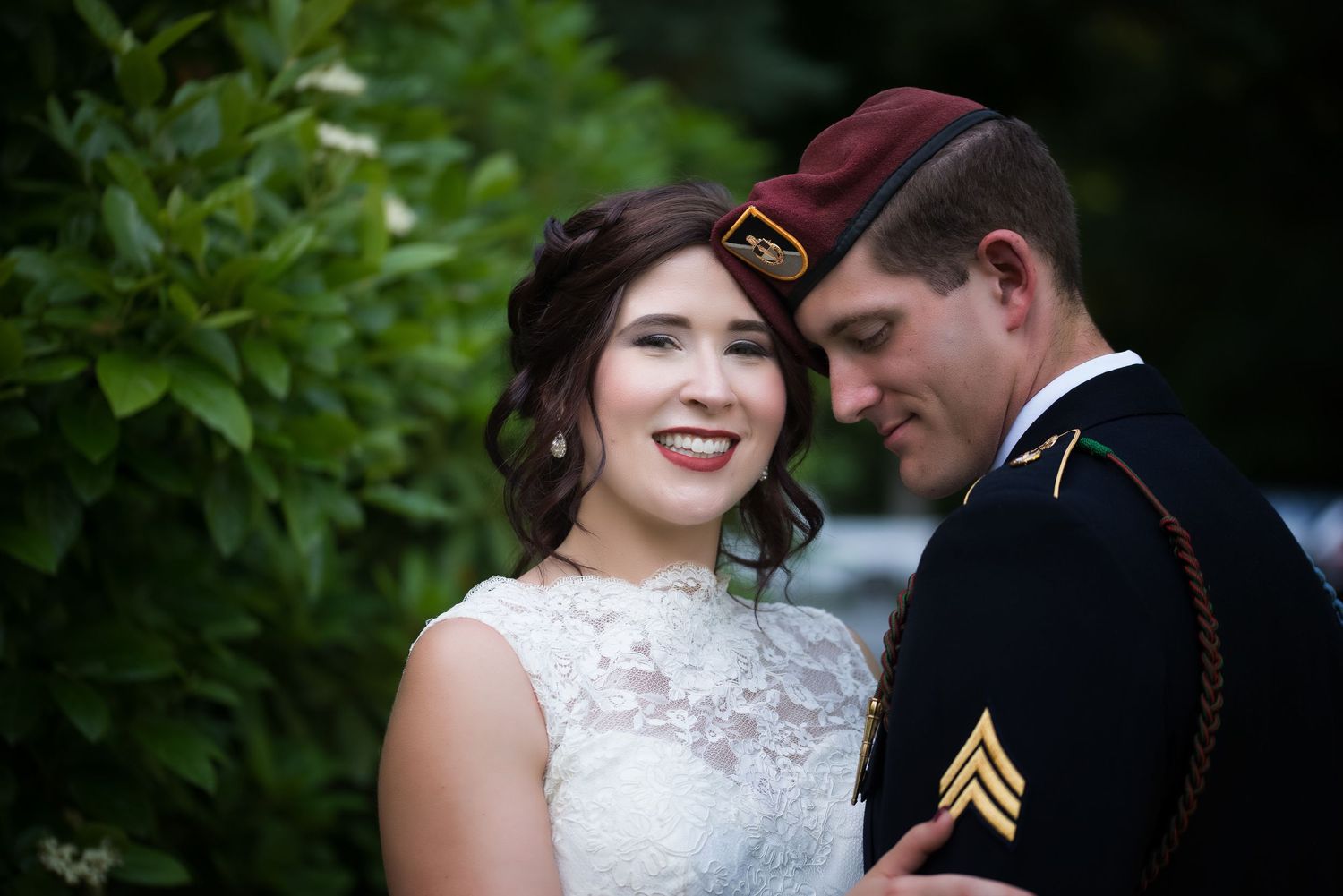 Smiling bride poses with groom in military dress uniform surrounded by lush green foliage at wedding.
