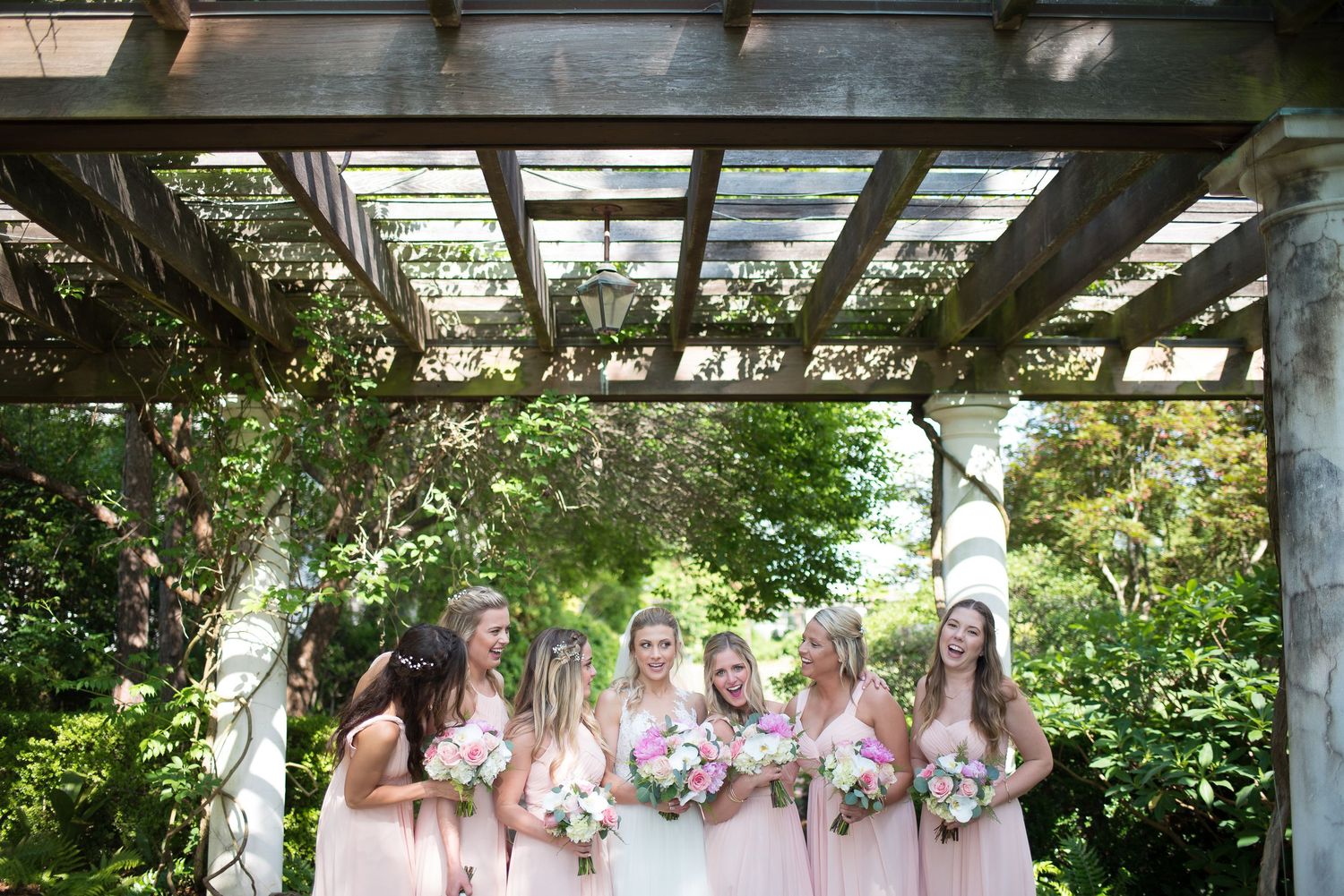 Bride and bridesmaids in blush pink gowns holding bouquets gather under a rustic wooden pergola outdoors.