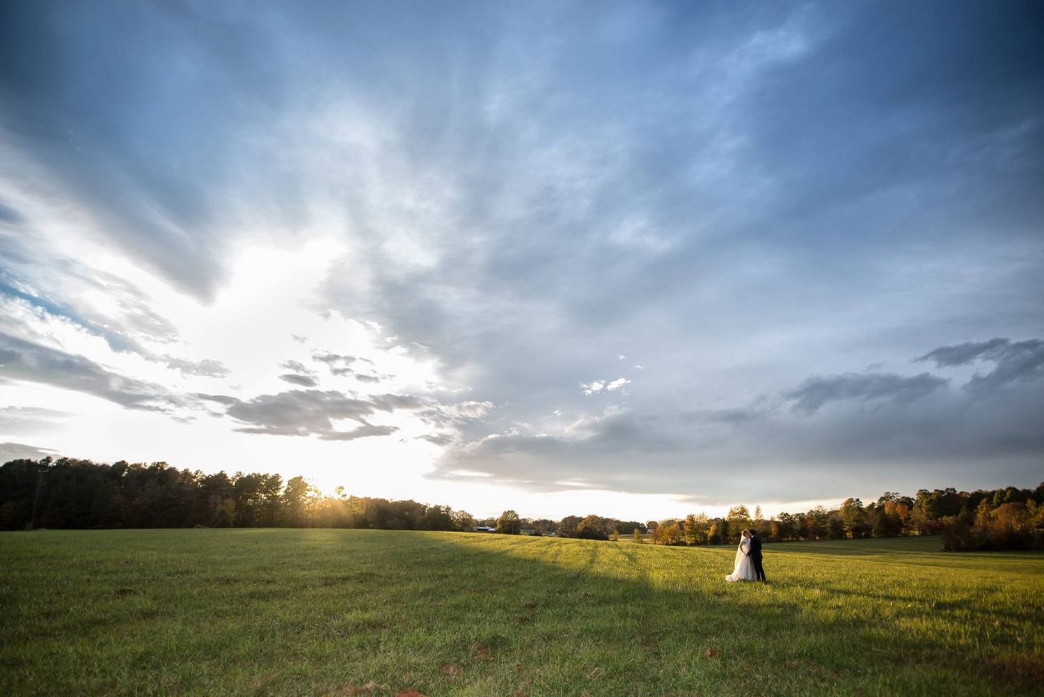 Bride and groom stand in a vast green field at sunset with dramatic cloudy sky backdrop.