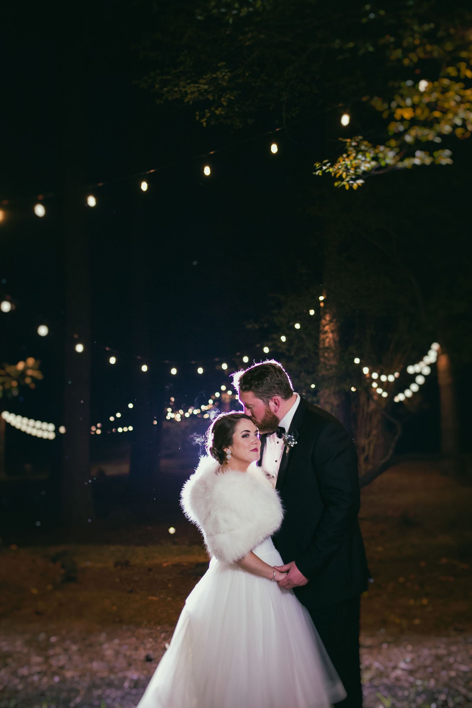 Bride and groom embracing under string lights in a romantic outdoor nighttime setting.