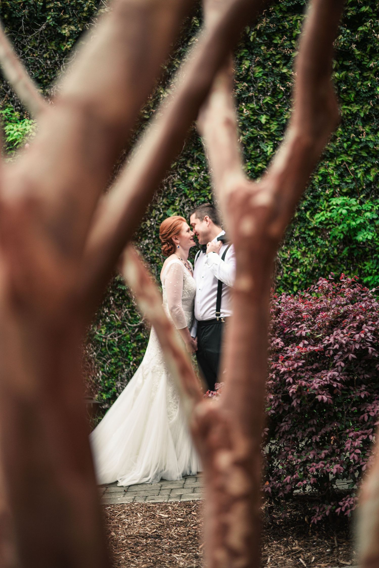 Bride and groom share a romantic kiss in a lush garden, framed by twisted branches and green hedges.