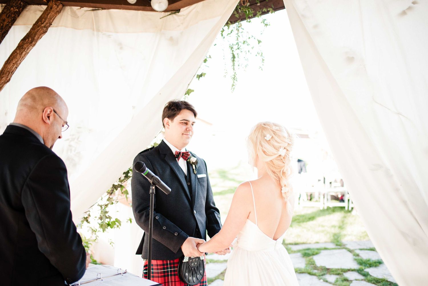 Bride and groom exchange vows at outdoor wedding ceremony under white draped canopy with officiant in tartan kilt.